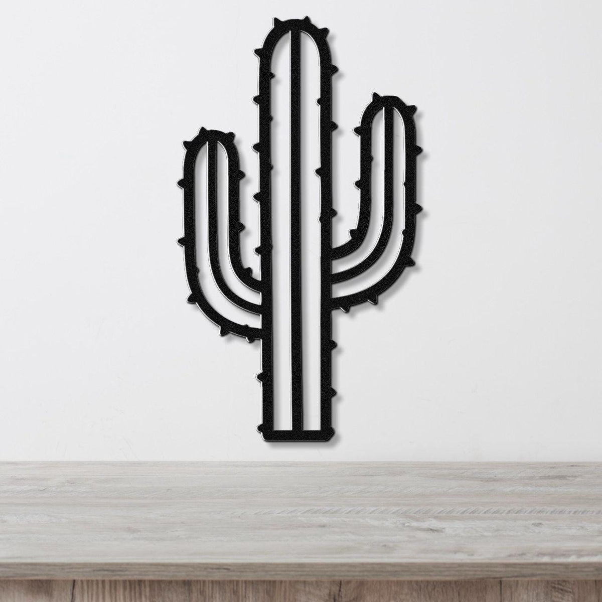 Black metal wall art in the silhouette of a saguaro cactus with spines and side arms, mounted on a white wall above a light wood surface.