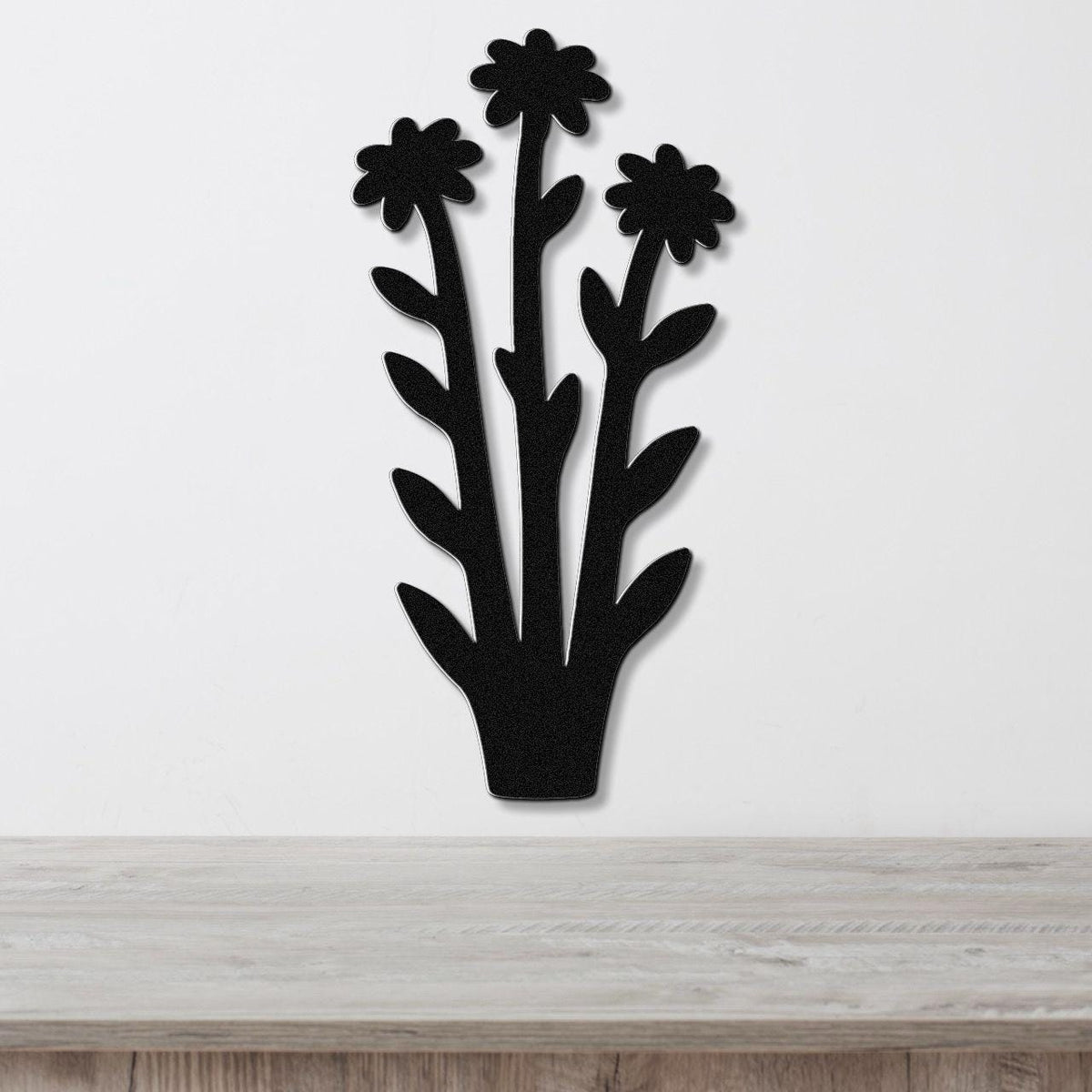 Black metal wall art featuring a silhouette of three wildflowers with leaves, mounted on a white wall above a wooden surface.