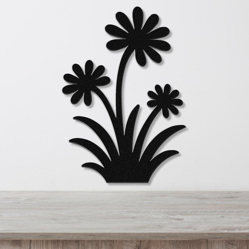 Black metal wall art featuring a silhouette of three daisy-like flowers with long stems and leafy base, mounted on a white wall above a light wood surface.