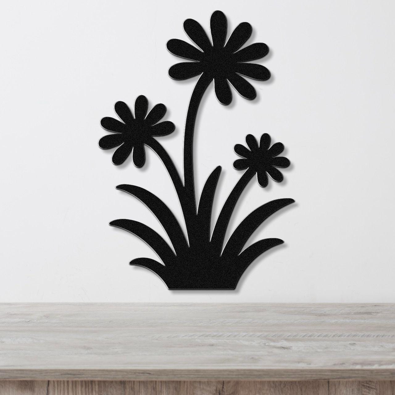 Black metal wall art featuring a silhouette of three daisy-like flowers with long stems and leafy base, mounted on a white wall above a light wood surface.