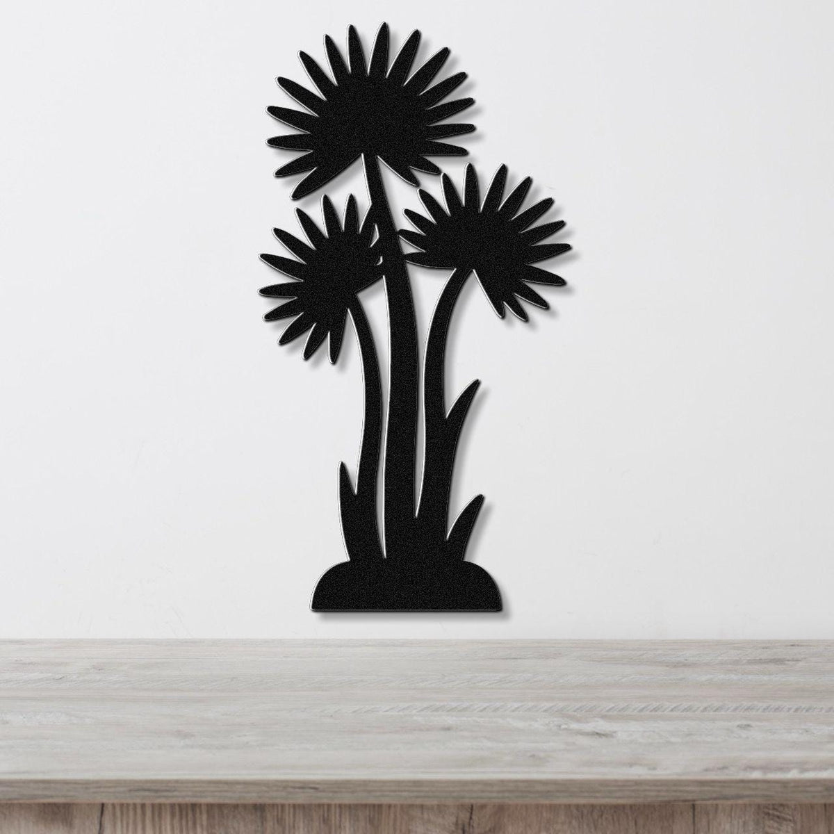 Black metal wall art featuring a silhouette of three wildflowers with radiating petals, mounted on a white wall above a wooden surface.
