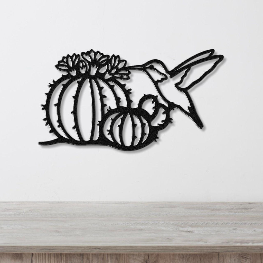 Black metal wall art featuring a hummingbird and barrel cactus with flower cutouts, mounted on a white wall above a wooden surface.