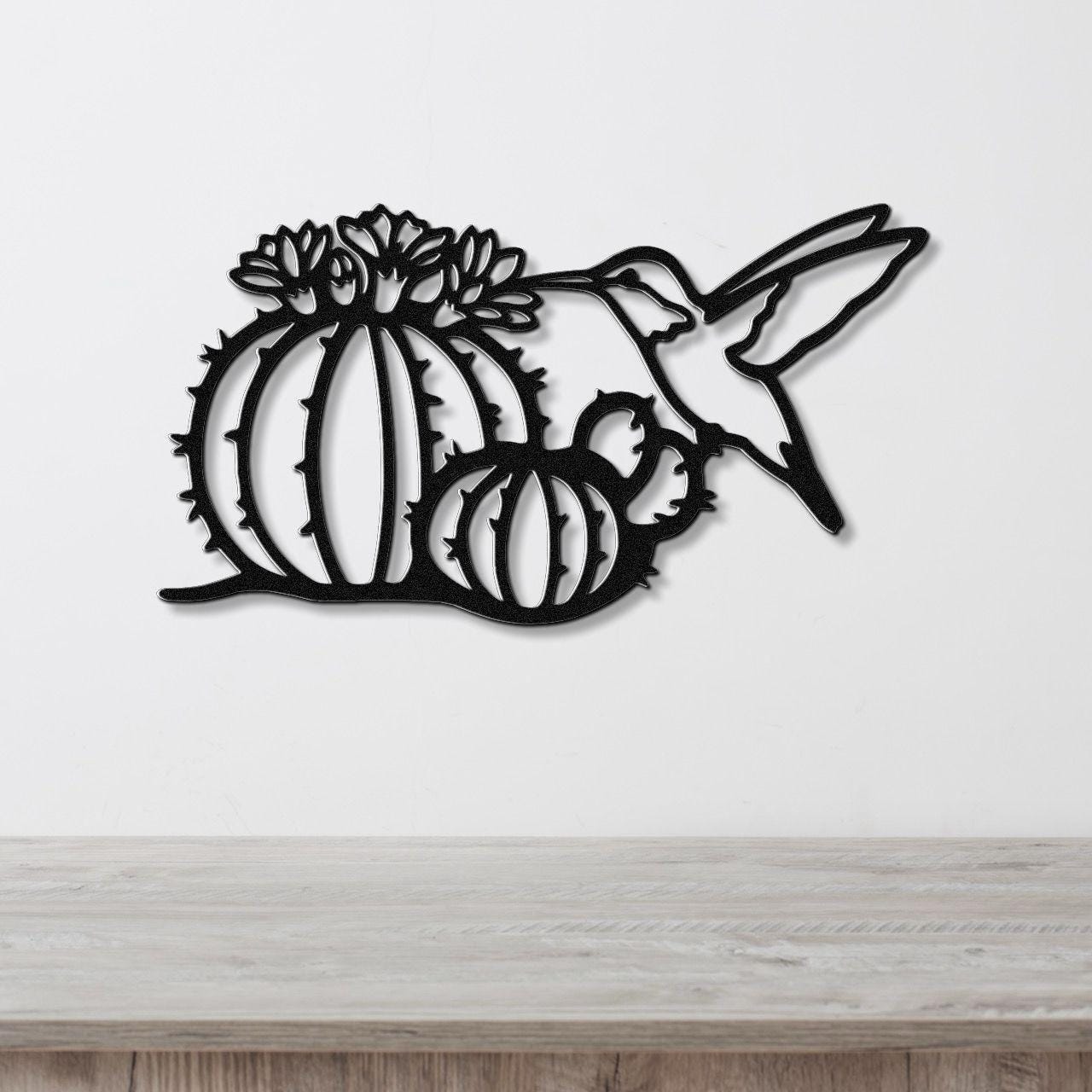 Black metal wall art featuring a hummingbird and barrel cactus with flower cutouts, mounted on a white wall above a wooden surface.