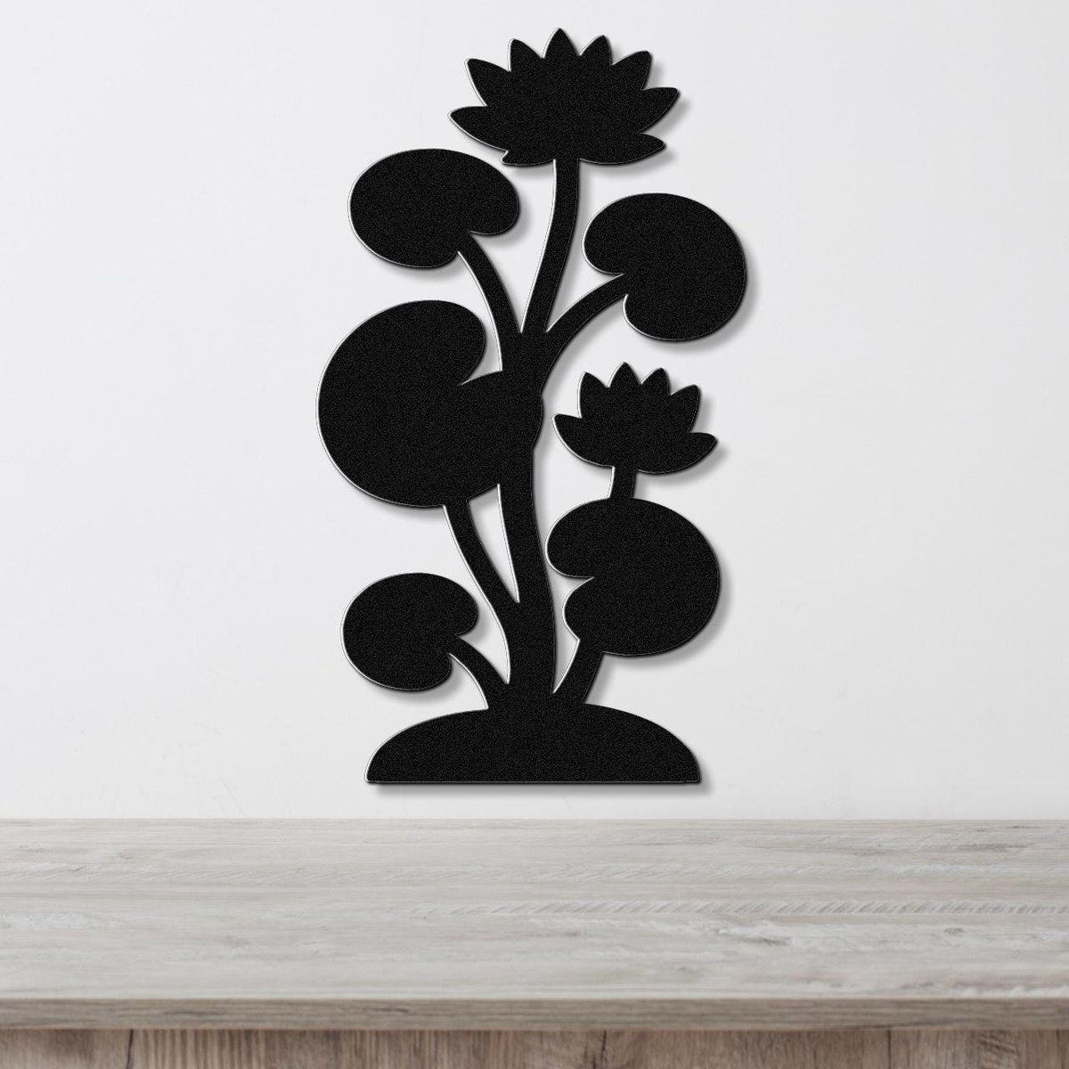 Black metal wall art cutout of a lotus plant with two flowers and lily pads, mounted on a white wall above a wooden surface.