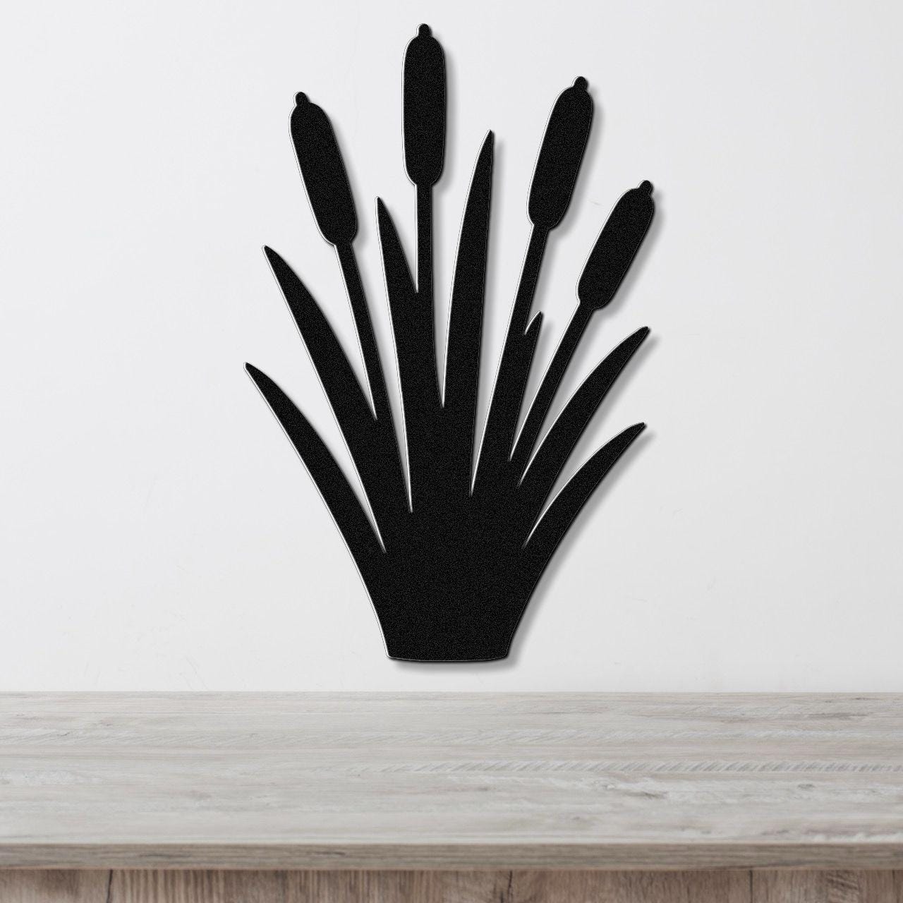 Black metal cattail wall art with a silhouette design of tall reeds and seed heads, mounted on a white wall above a light wood surface.