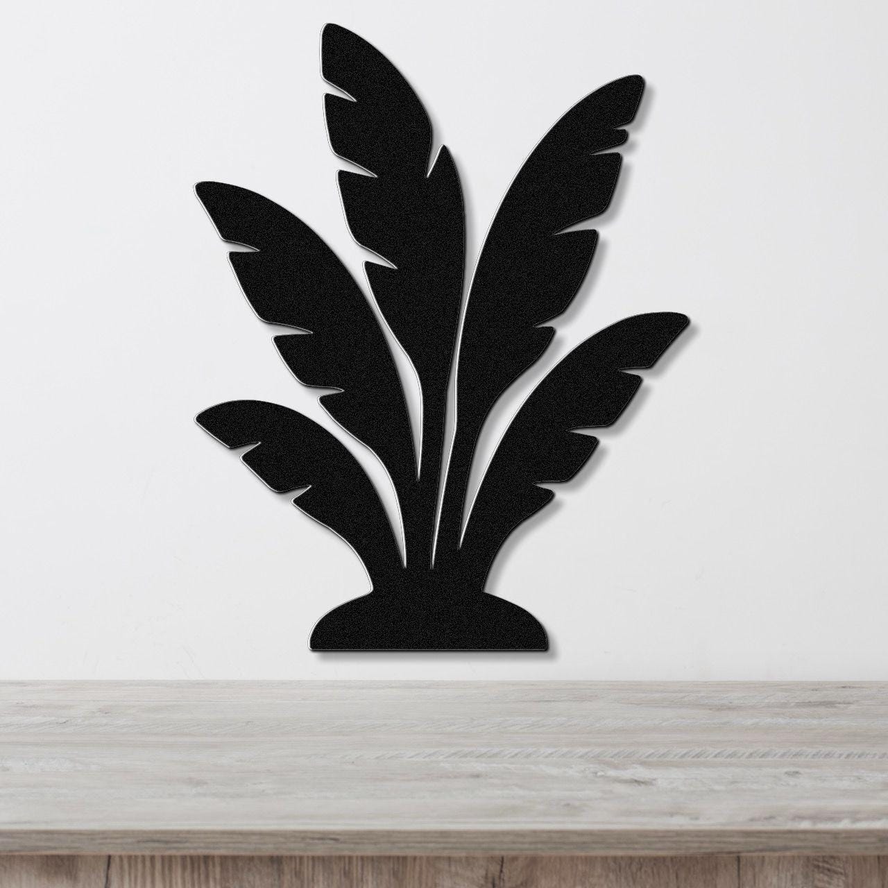 Black metal wall art in a tropical leaf silhouette design, mounted on a white wall above a light wood surface.
