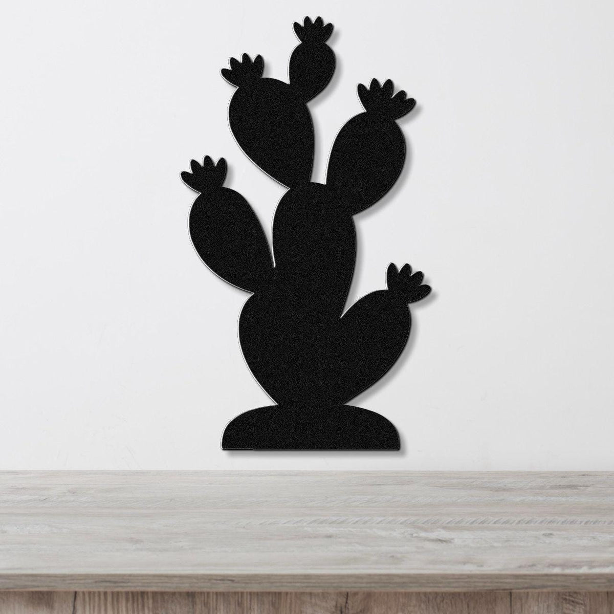 Black metal cactus wall art with multiple paddle-shaped arms and flower cutouts, mounted on a white wall above a light wood surface.