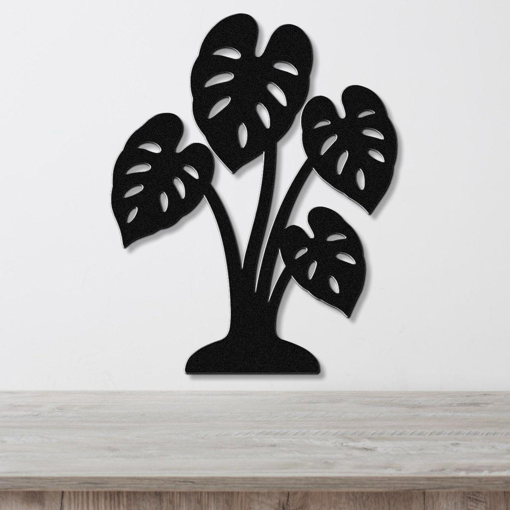 Black metal wall art cutout of a monstera plant with leaf perforations, mounted on a white wall above a wooden surface.