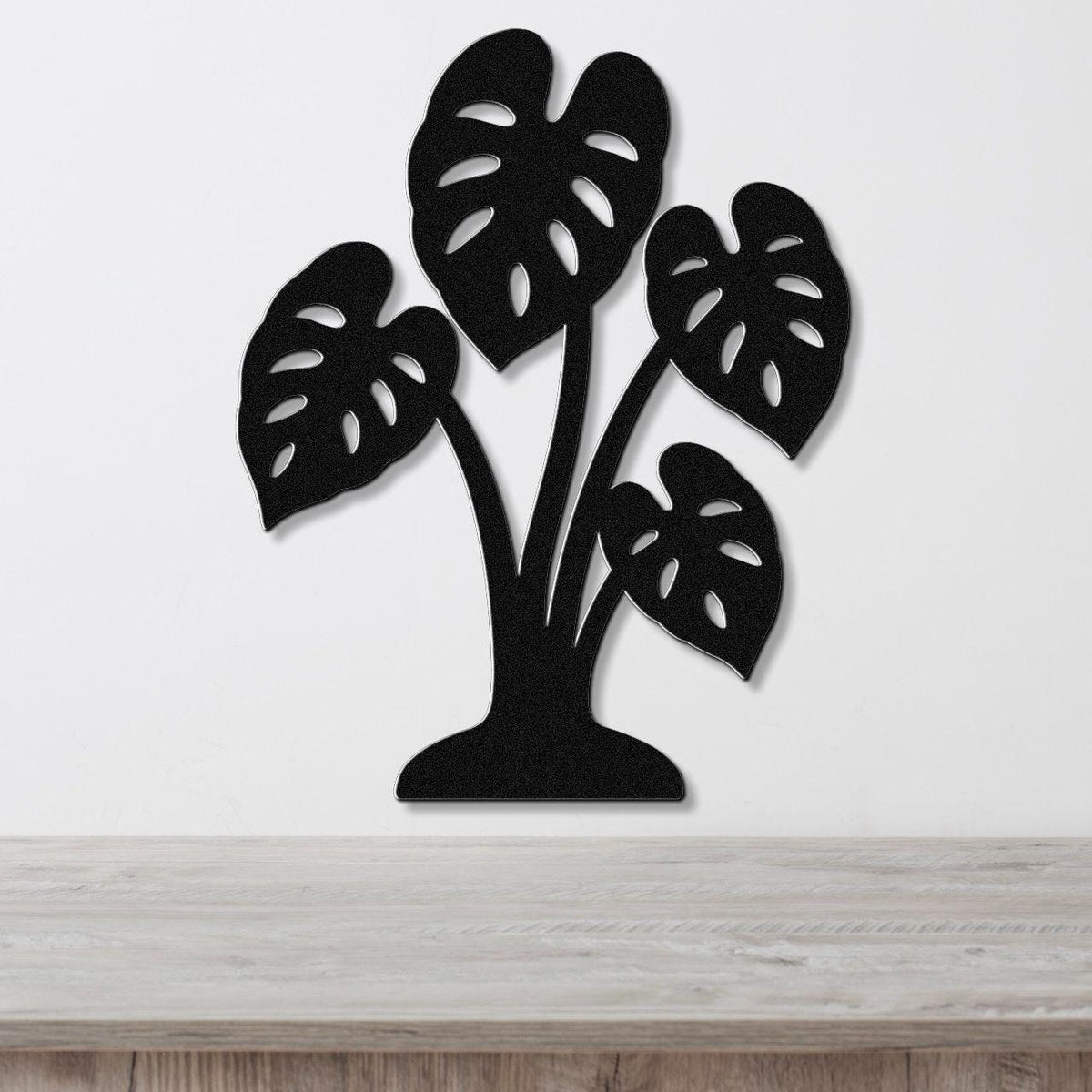 Black metal wall art cutout of a monstera plant with leaf perforations, mounted on a white wall above a wooden surface.