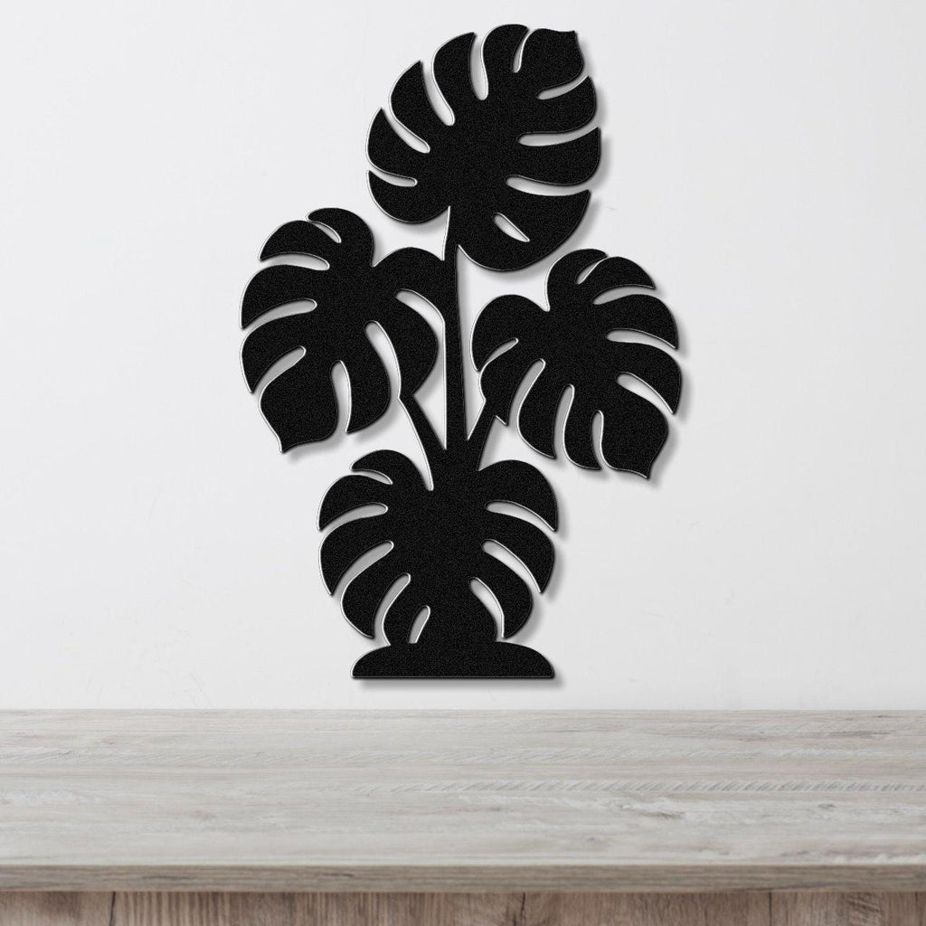 Black metal wall art cutout of a monstera plant with multiple leaves, mounted on a white wall above a wooden surface.