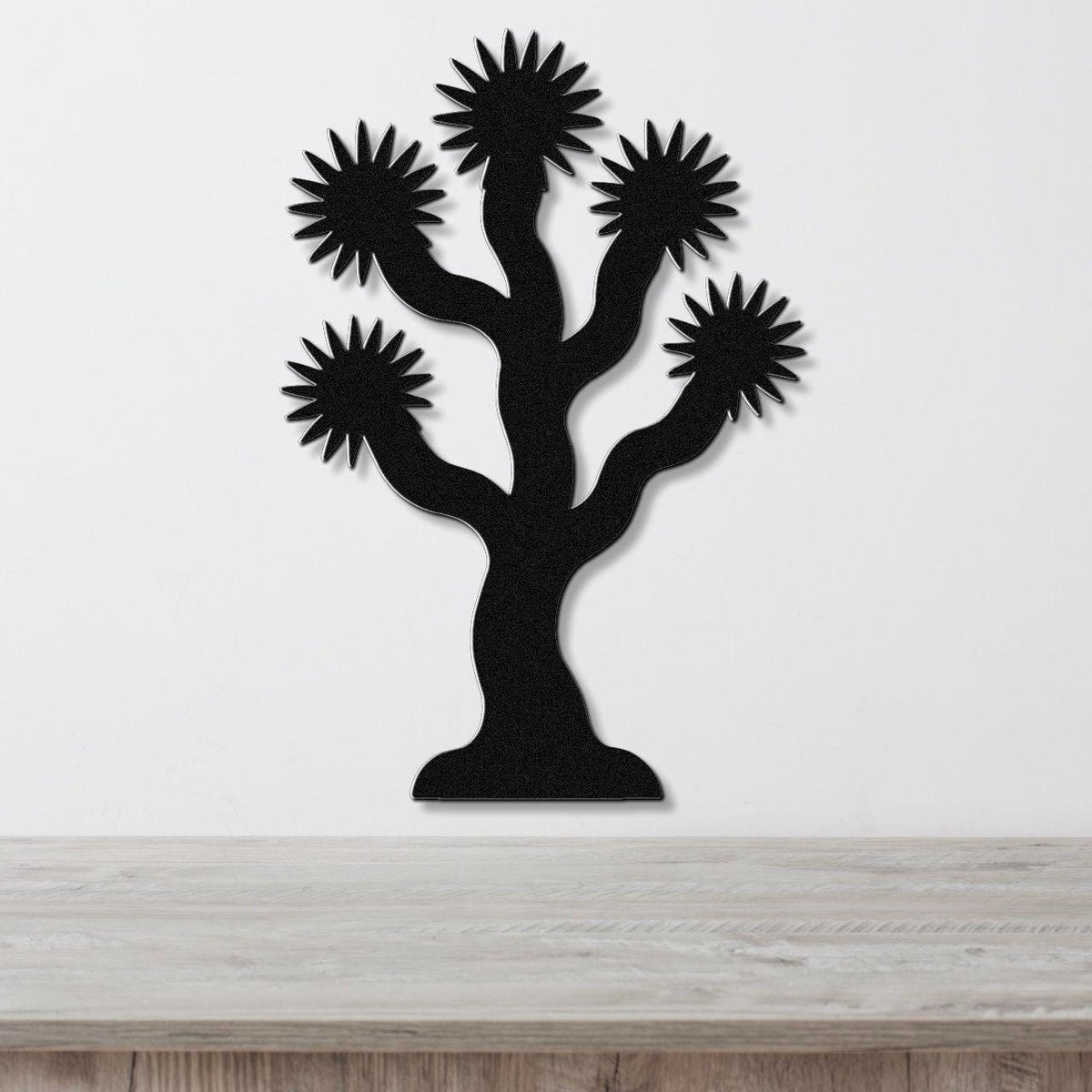 Black metal wall art in the silhouette of a Joshua tree with starburst-shaped branches, mounted on a white wall above a light wood surface.