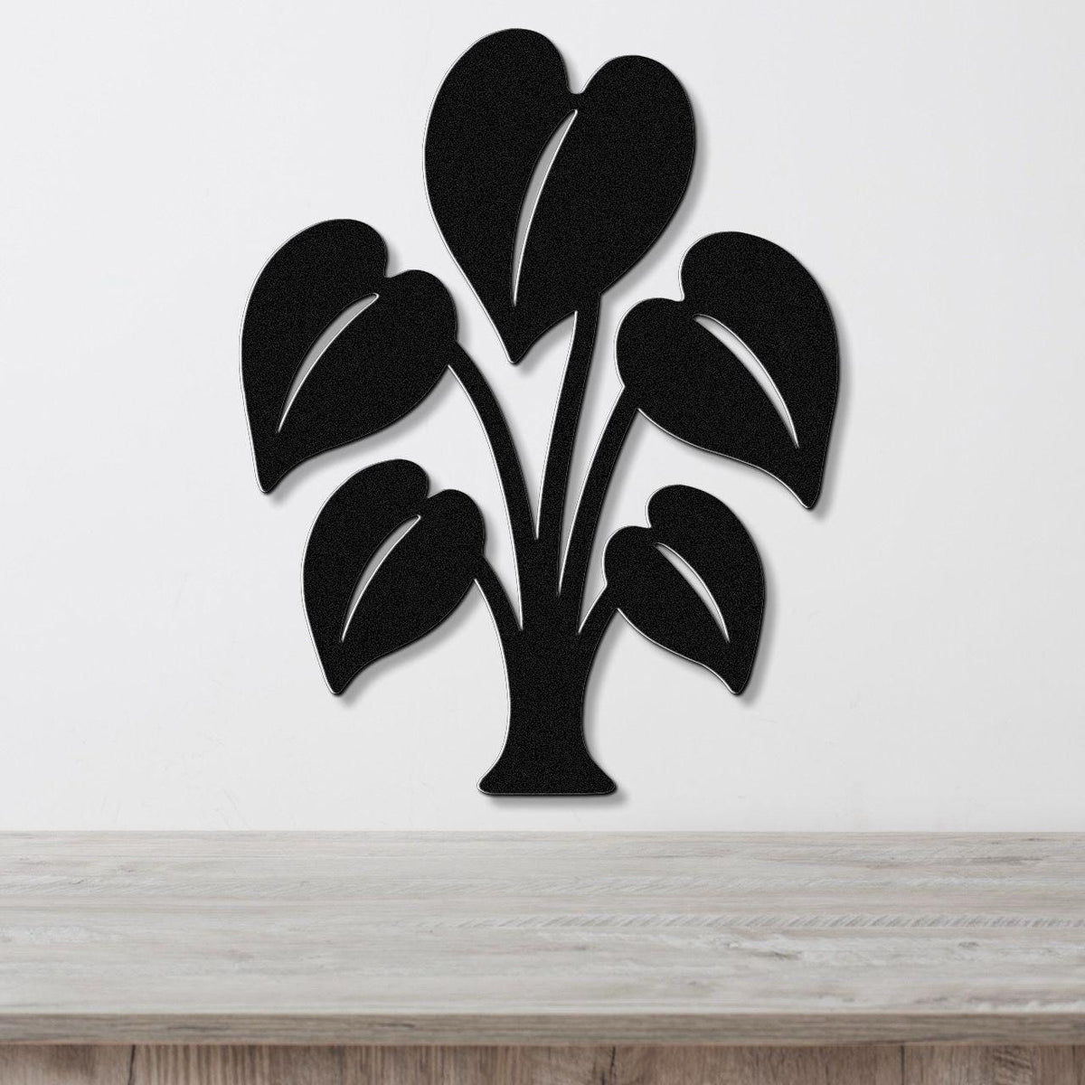 Black metal wall art in the shape of an elephant ear plant with leaf cutouts, mounted on a white wall above a wooden surface.