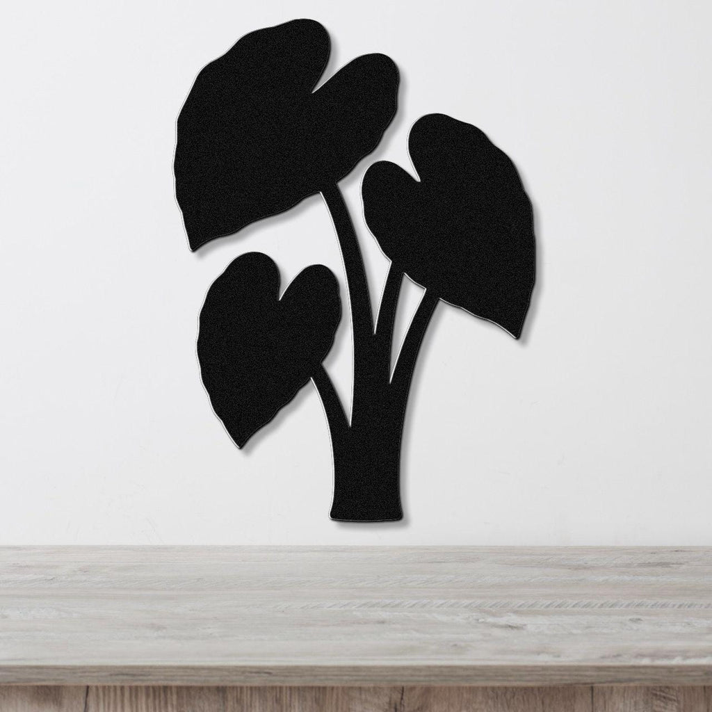 Black metal wall art in the silhouette of an elephant ear plant with three large leaves, mounted on a white wall above a light wood surface.