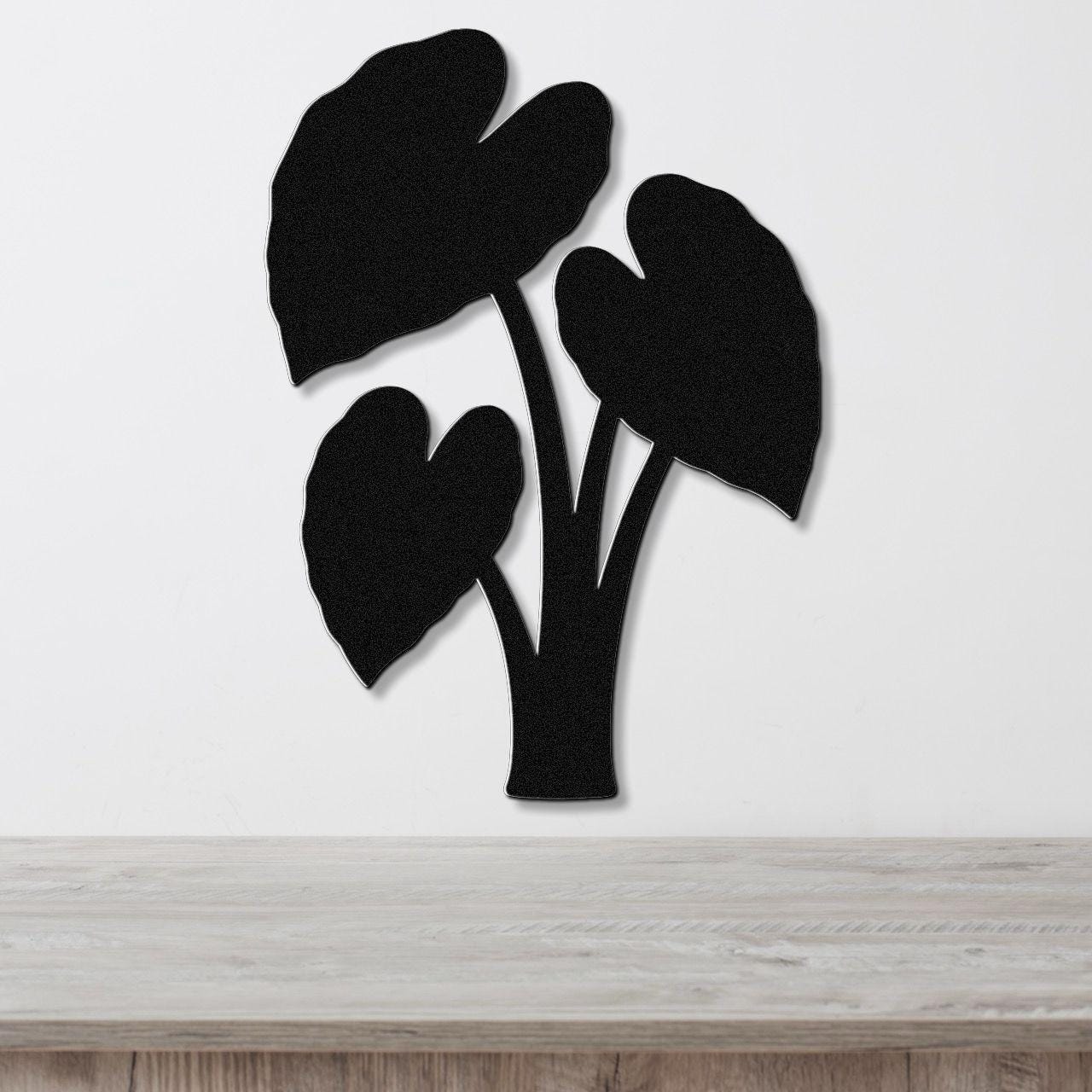 Black metal wall art in the silhouette of an elephant ear plant with three large leaves, mounted on a white wall above a light wood surface.