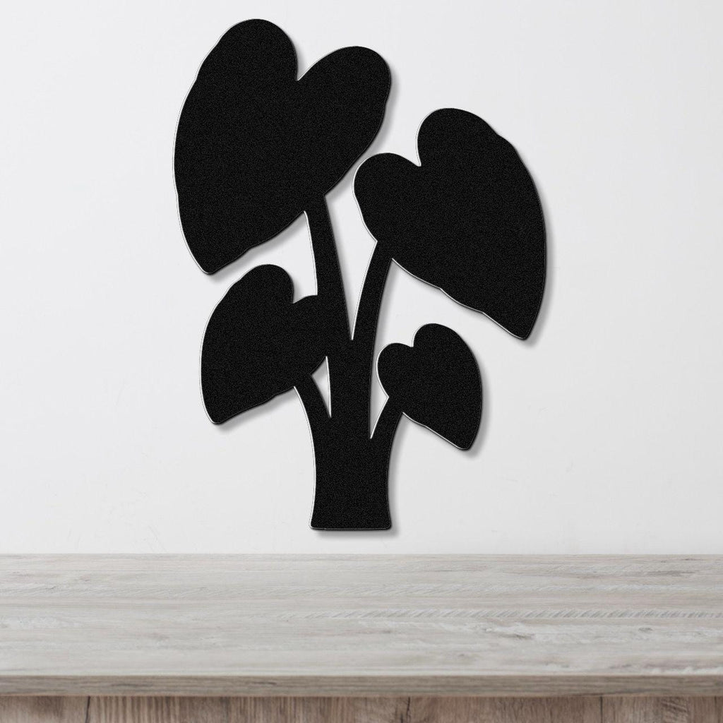 Black metal wall art in the shape of an elephant ear plant with heart-shaped leaves, mounted on a white wall above a wooden surface.