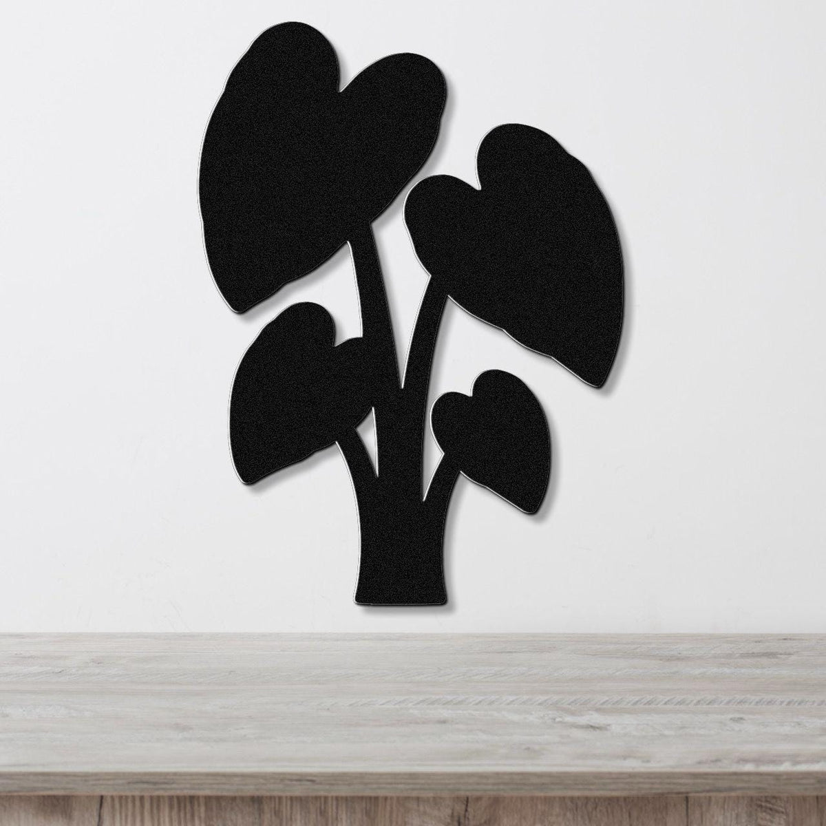 Black metal wall art in the shape of an elephant ear plant with heart-shaped leaves, mounted on a white wall above a wooden surface.