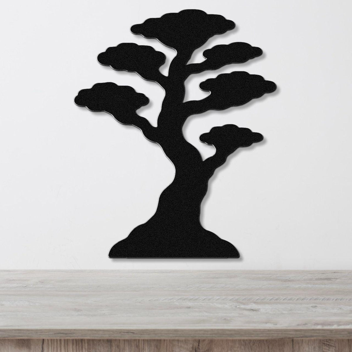 Black metal wall art in the silhouette of a bonsai tree with layered cloud-like branches, mounted on a white wall above a wooden surface.