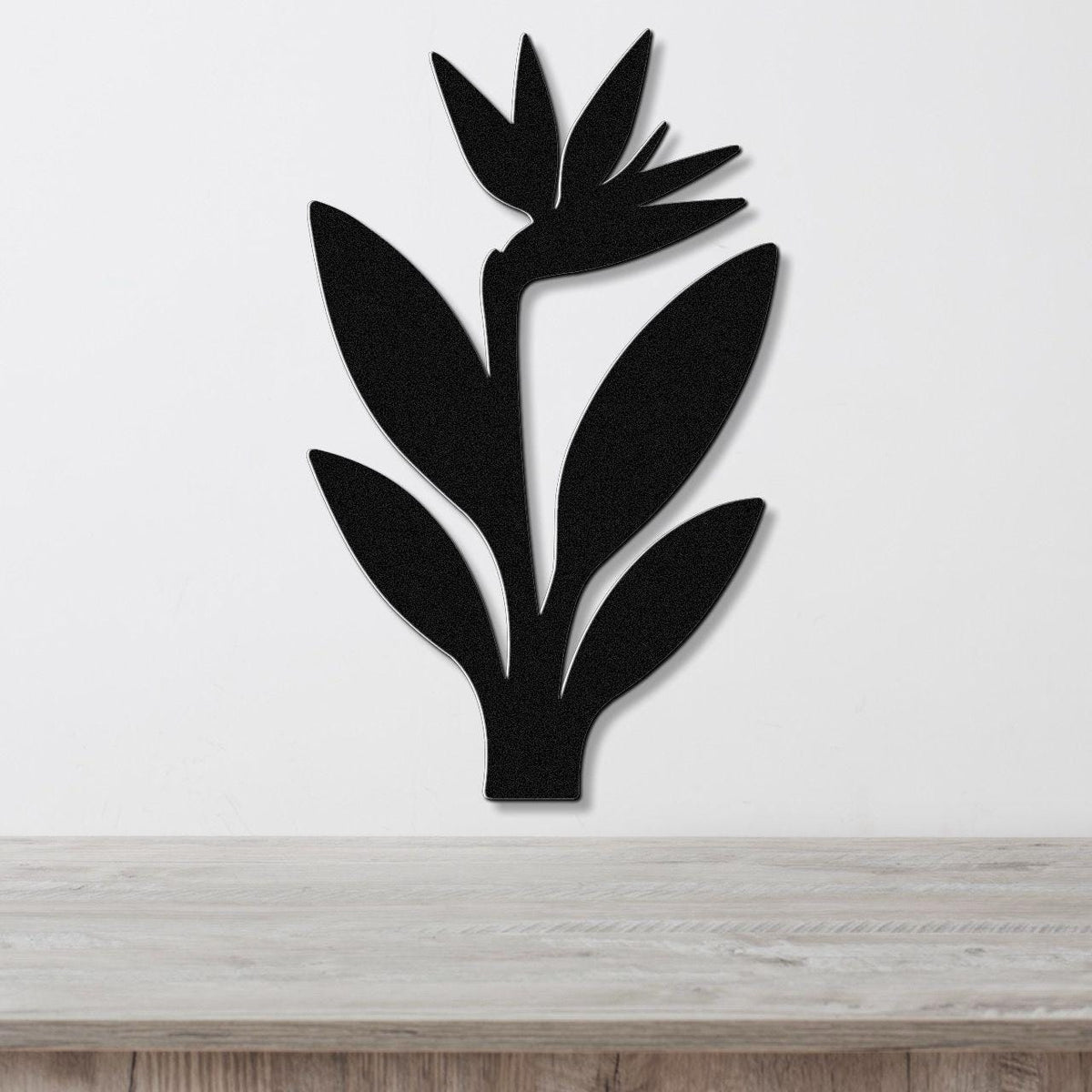 Black metal wall art in a clean Bird of Paradise silhouette with layered leaves and flower cutouts, mounted on a white wall above a light wood surface.