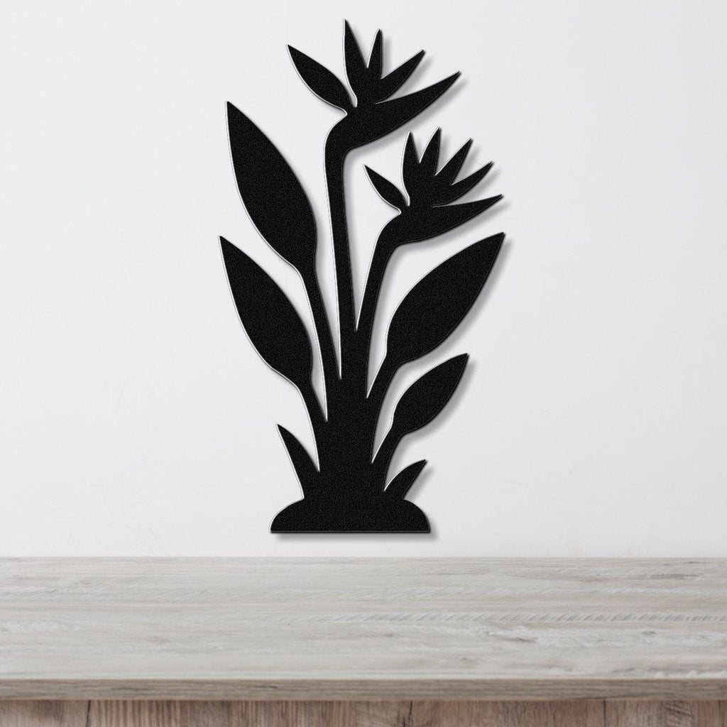 Black metal wall art silhouette of a Bird of Paradise plant with layered leaves and flowers, mounted on a white wall above a light wood surface.