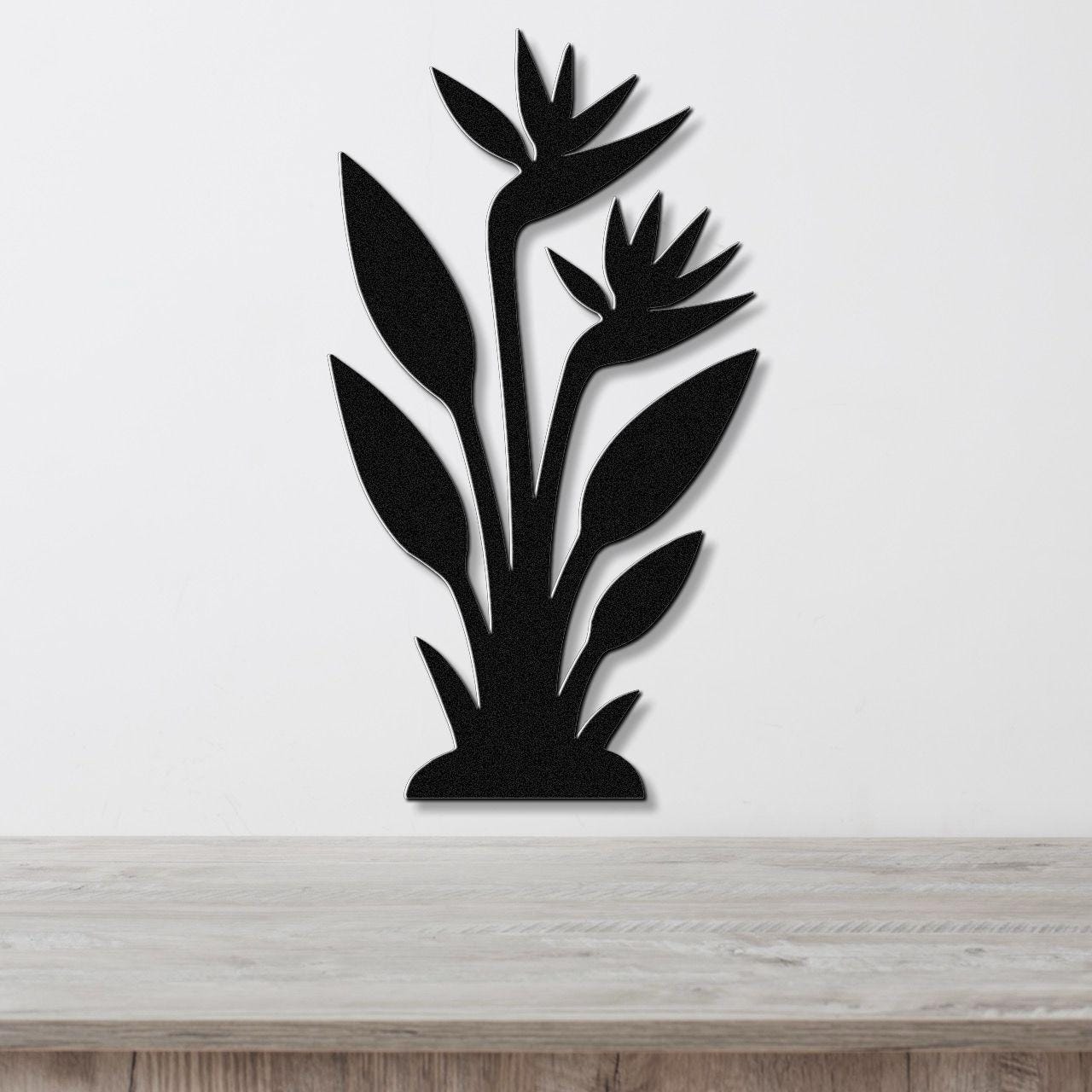 Black metal wall art silhouette of a Bird of Paradise plant with layered leaves and flowers, mounted on a white wall above a light wood surface.