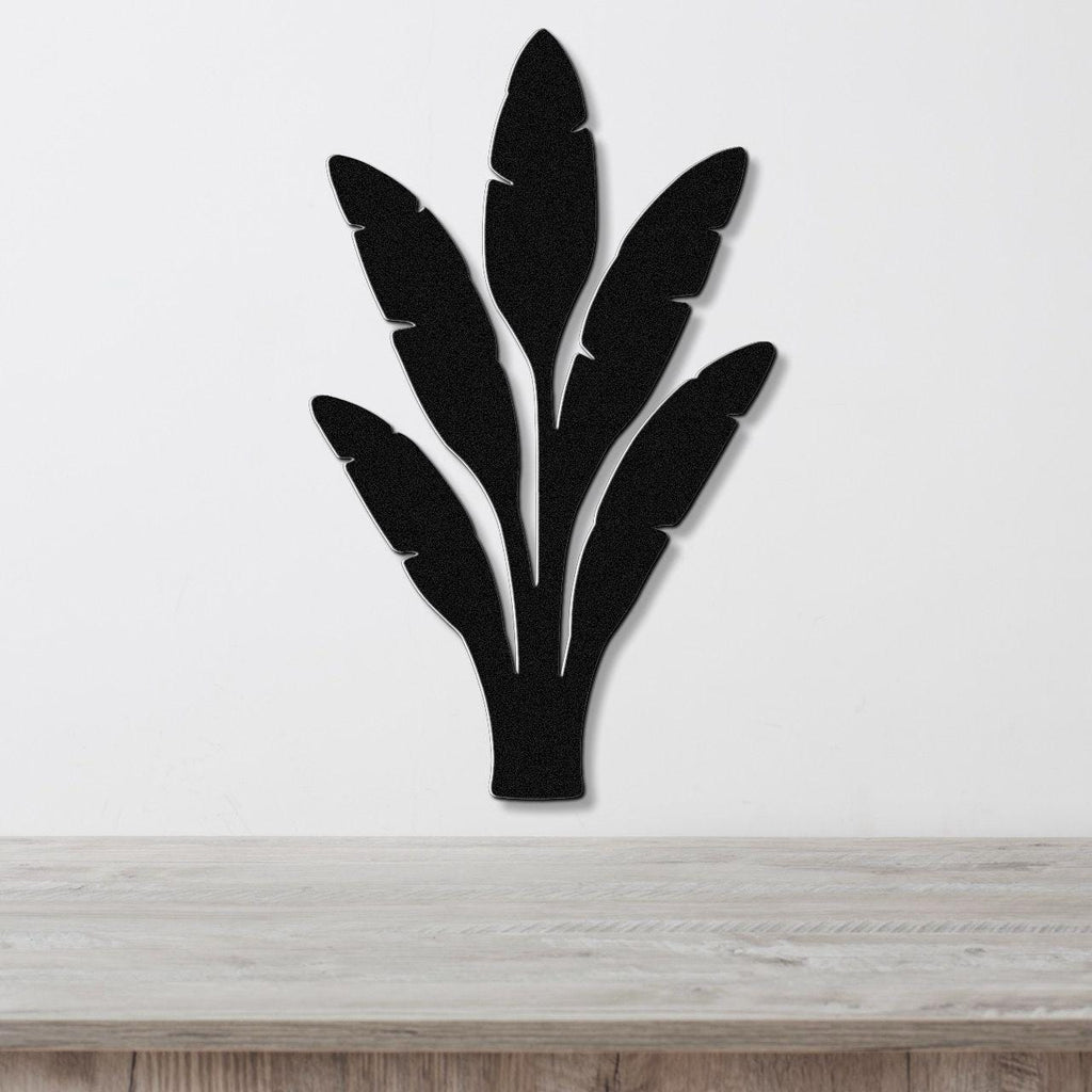 Black metal wall art in a banana leaf silhouette with layered cutouts, mounted on a white wall above a light wood counter.