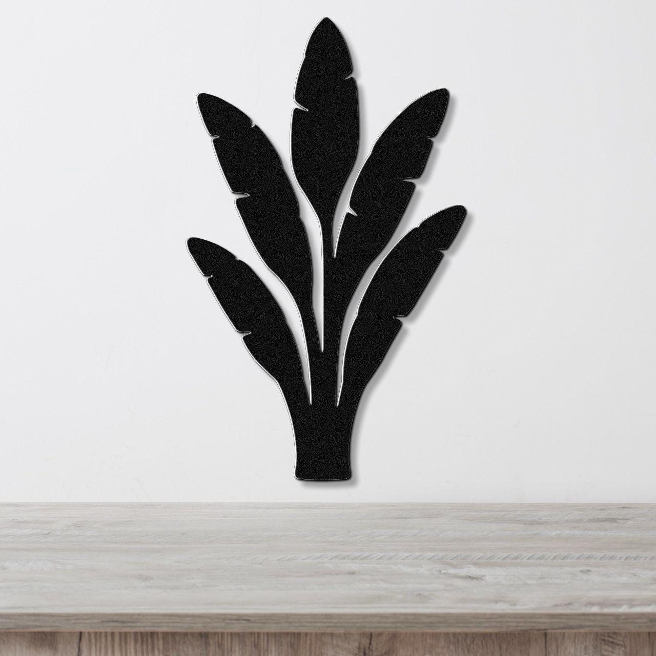 Black metal wall art in a banana leaf silhouette with layered cutouts, mounted on a white wall above a light wood counter.
