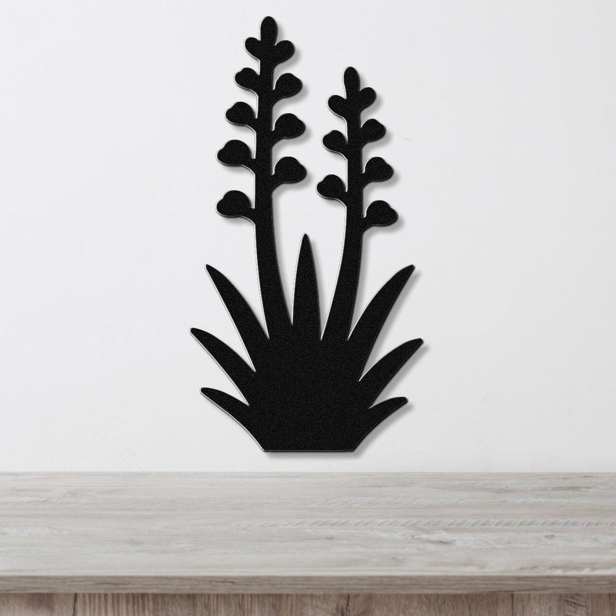 Black metal wall art cutout of a blooming agave plant with two tall flower stalks and spiky base, mounted on a white wall above a light wood surface.