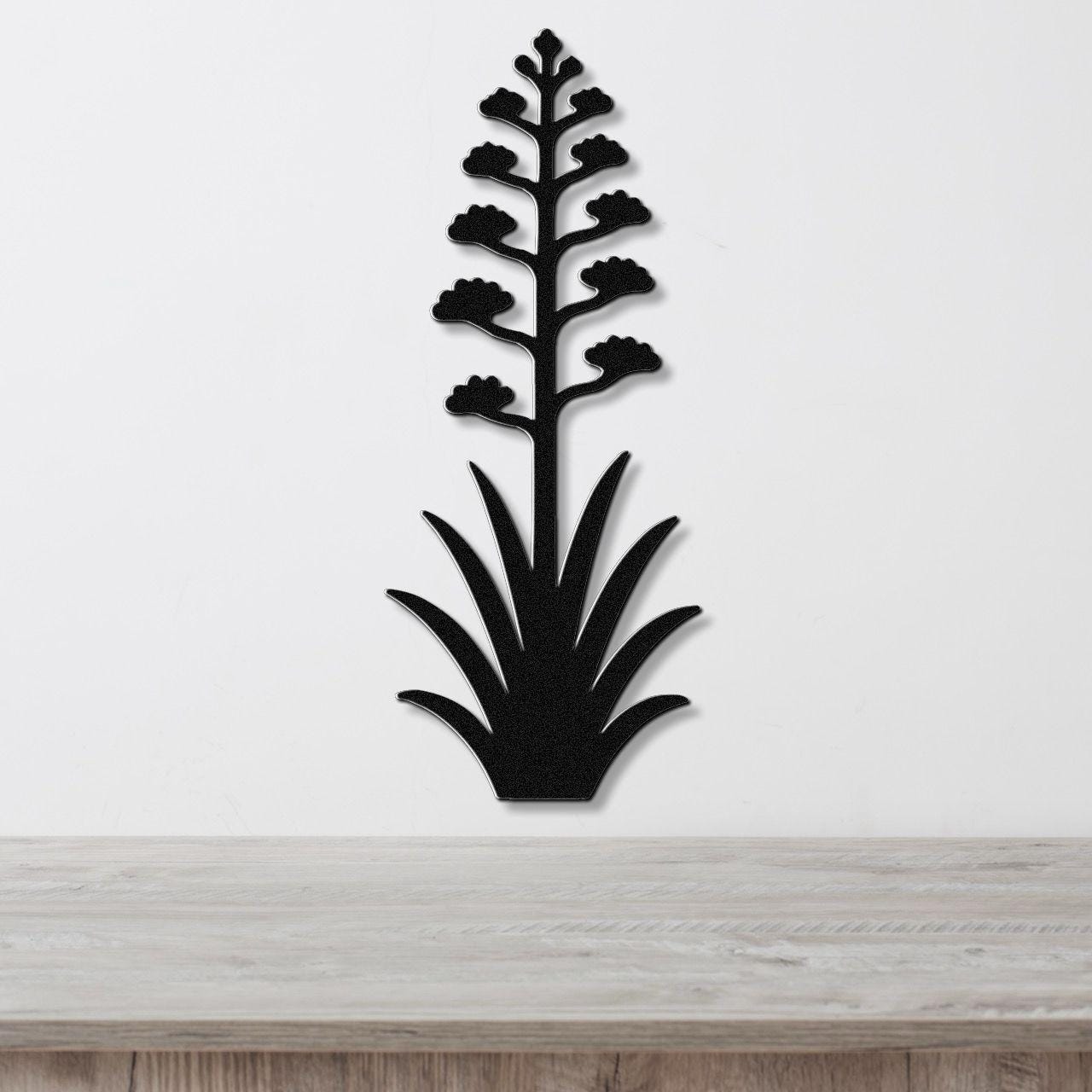 Black metal wall art in the silhouette of a blooming agave plant with a tall stem and spiky leaves, mounted on a white wall above a wooden surface.