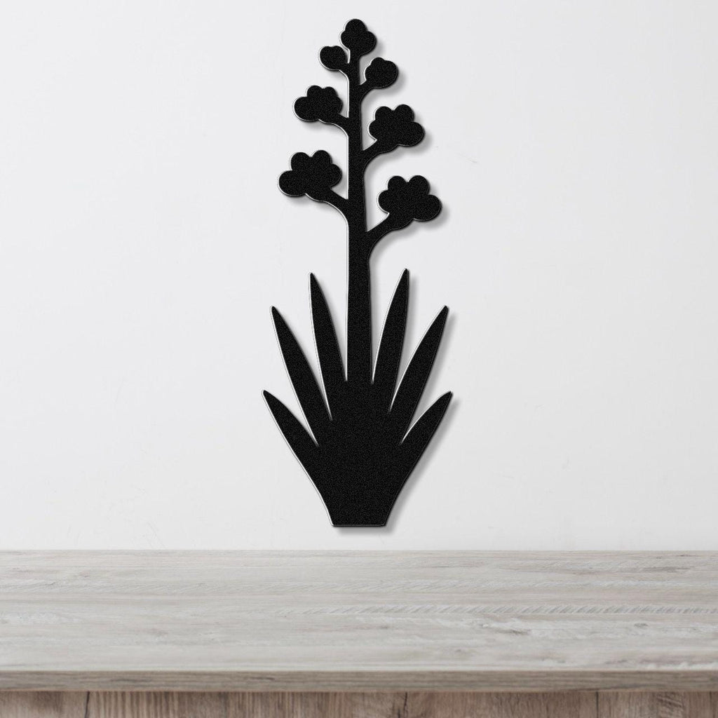 Black metal wall art silhouette of a blooming agave plant with spiky leaves and flower clusters, mounted on a white wall above a light wood surface.
