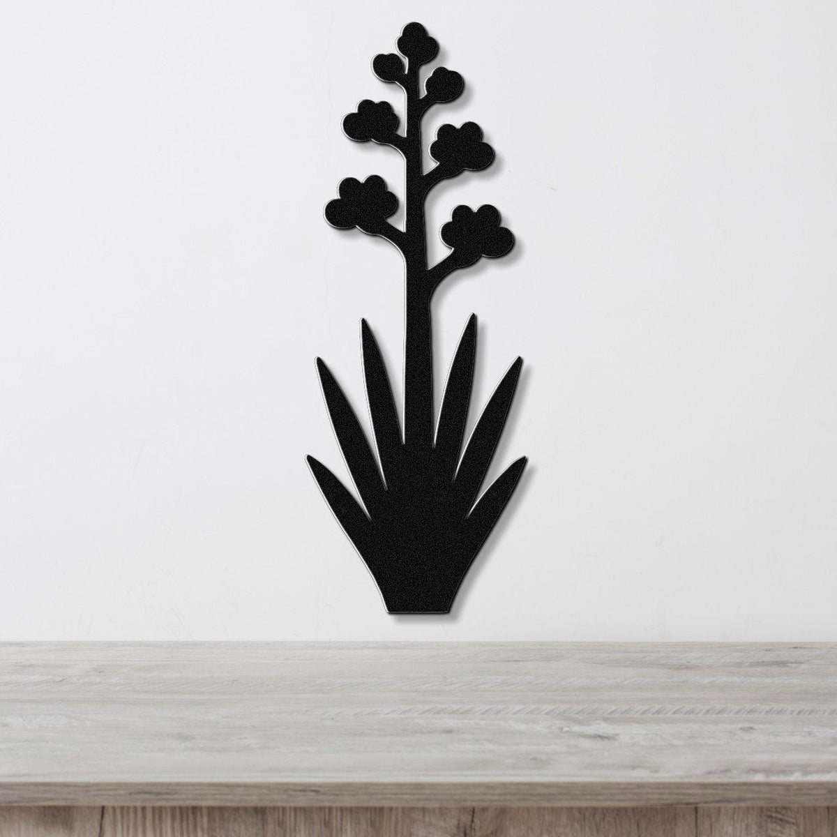 Black metal wall art silhouette of a blooming agave plant with spiky leaves and flower clusters, mounted on a white wall above a light wood surface.