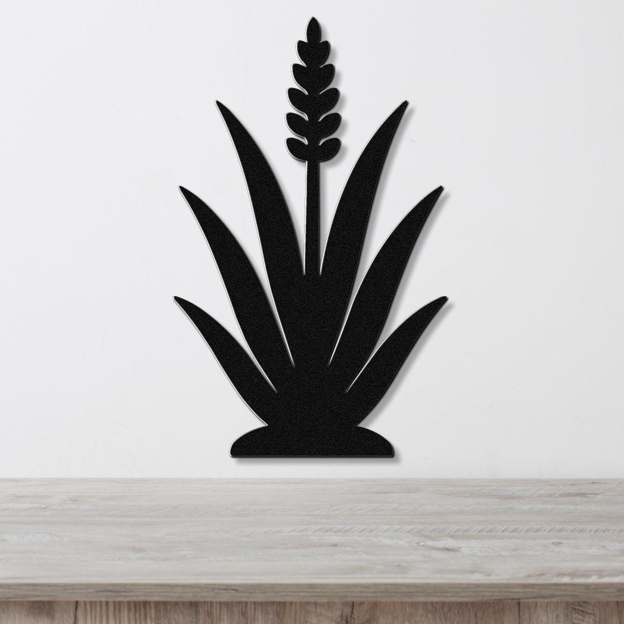 Black metal agave wall art with a central flower spike and leaf cutouts, mounted on a white wall above a light wood surface.