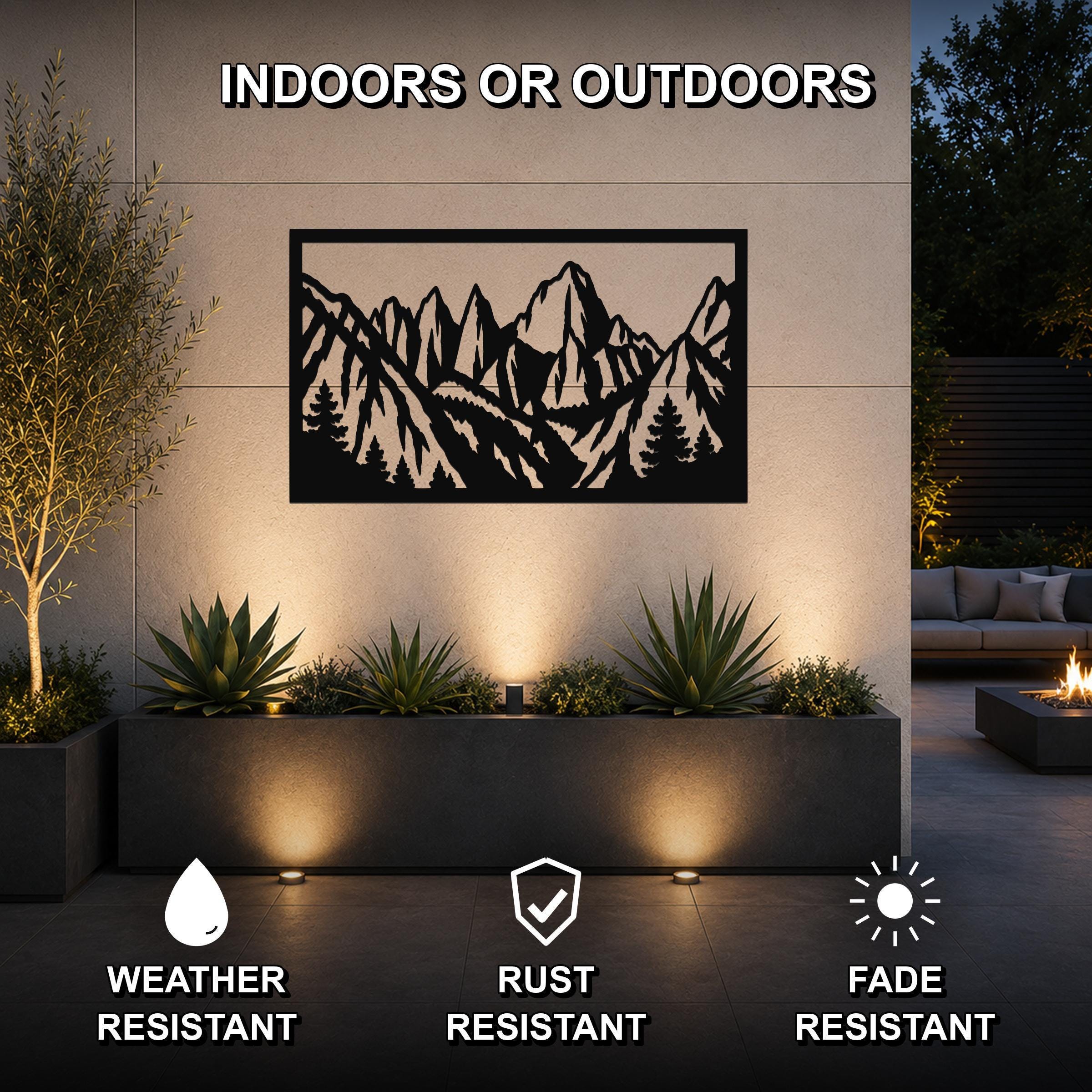 Black metal wall art with a mountain and tree silhouette cutout, mounted on an outdoor wall above a planter in a patio setting.