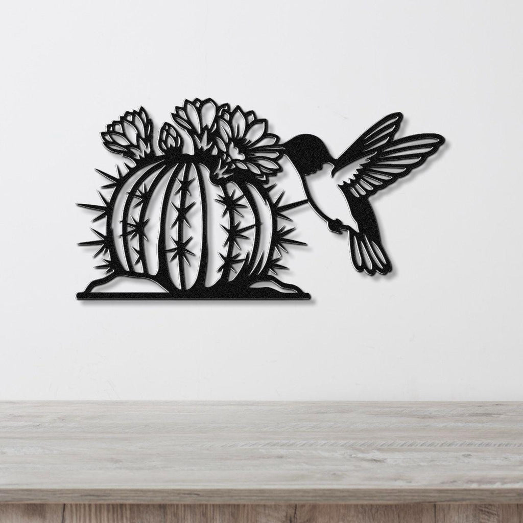 Black metal wall art featuring a hummingbird in flight approaching a flowering barrel cactus with detailed spines and cutout flowers, mounted on a white wall above a wooden surface.