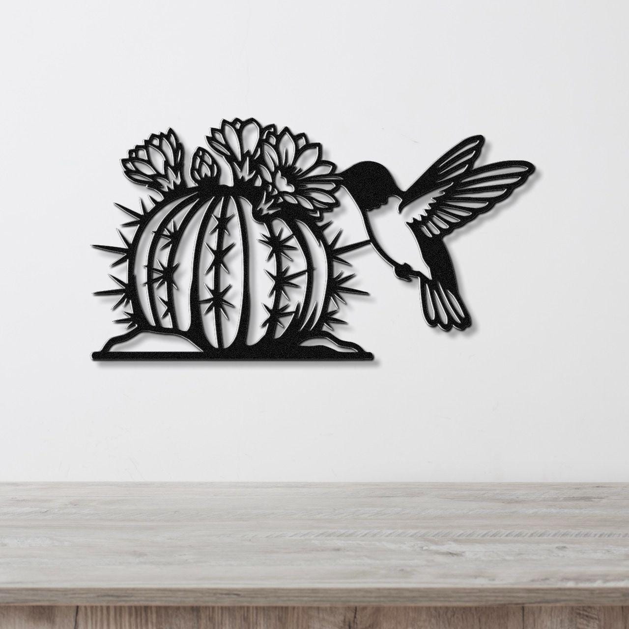 Black metal wall art featuring a hummingbird in flight approaching a flowering barrel cactus with detailed spines and cutout flowers, mounted on a white wall above a wooden surface.