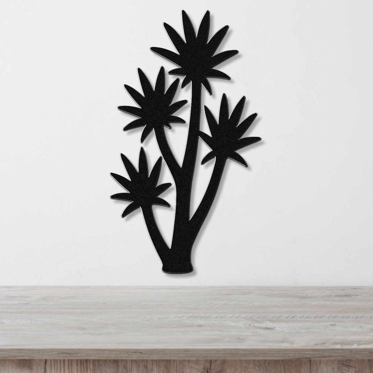 Black metal wall art cutout of a tall yucca plant with multiple star-shaped fronds, mounted on a white wall above a light wood surface.