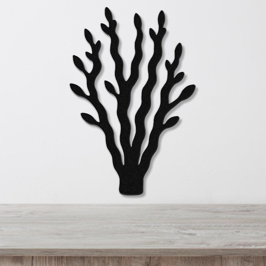 Black metal wall art in a kelp or seaweed silhouette with multiple branching fronds, mounted on a white wall above a light wood surface.