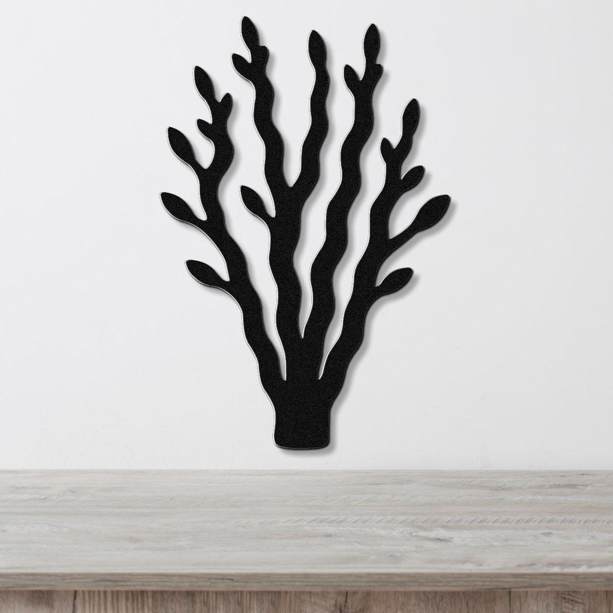 Black metal wall art in a kelp or seaweed silhouette with multiple branching fronds, mounted on a white wall above a light wood surface.