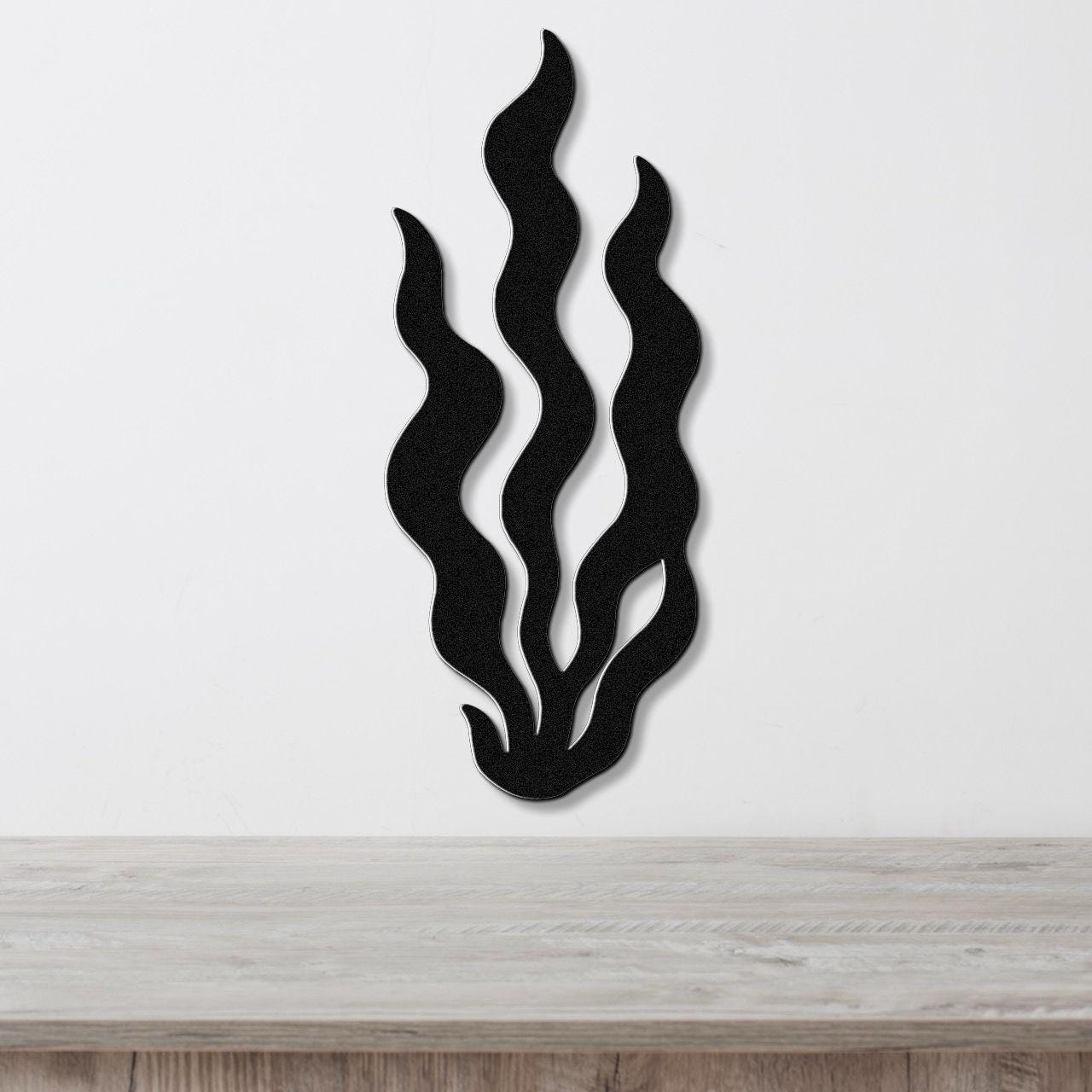 Black metal wall art in a kelp or seaweed silhouette design with flowing, wavy cutouts, mounted on a white wall above a light wood surface.