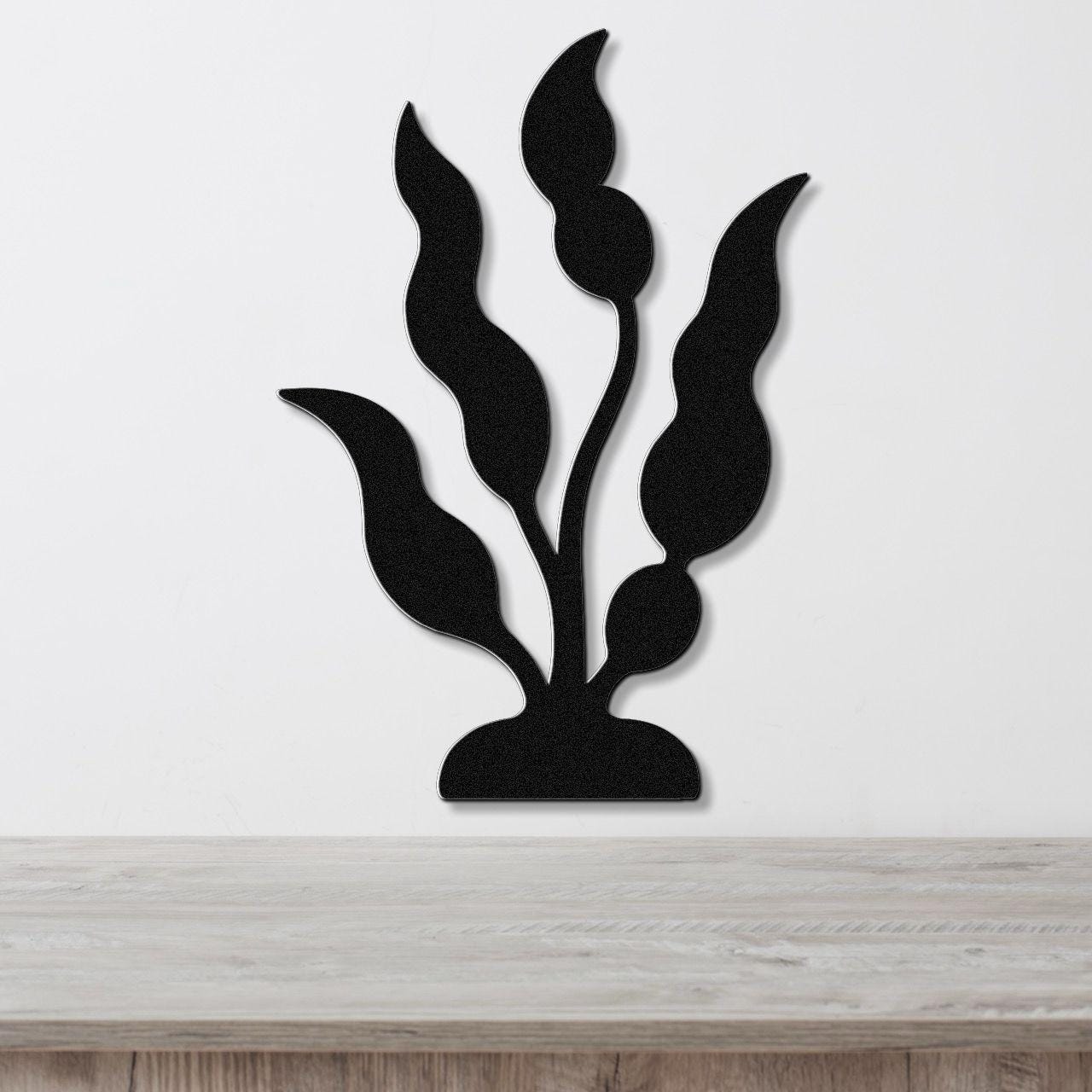 Black metal wall art cutout of kelp and seaweed with flowing leaf shapes, mounted on a white wall above a light wood surface.