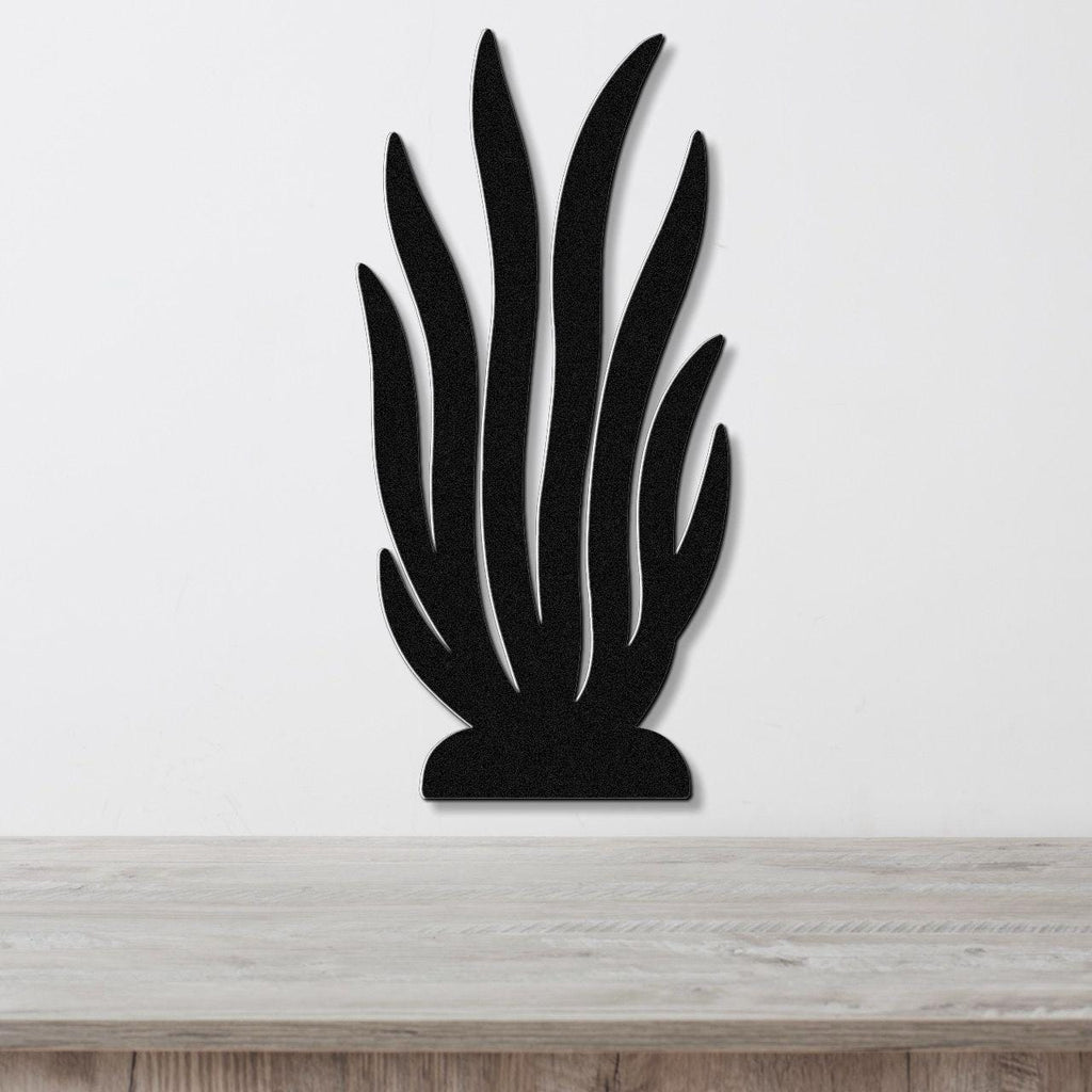 Black metal wall art in a kelp or seaweed silhouette design with multiple vertical fronds, mounted on a white wall above a light wood surface.