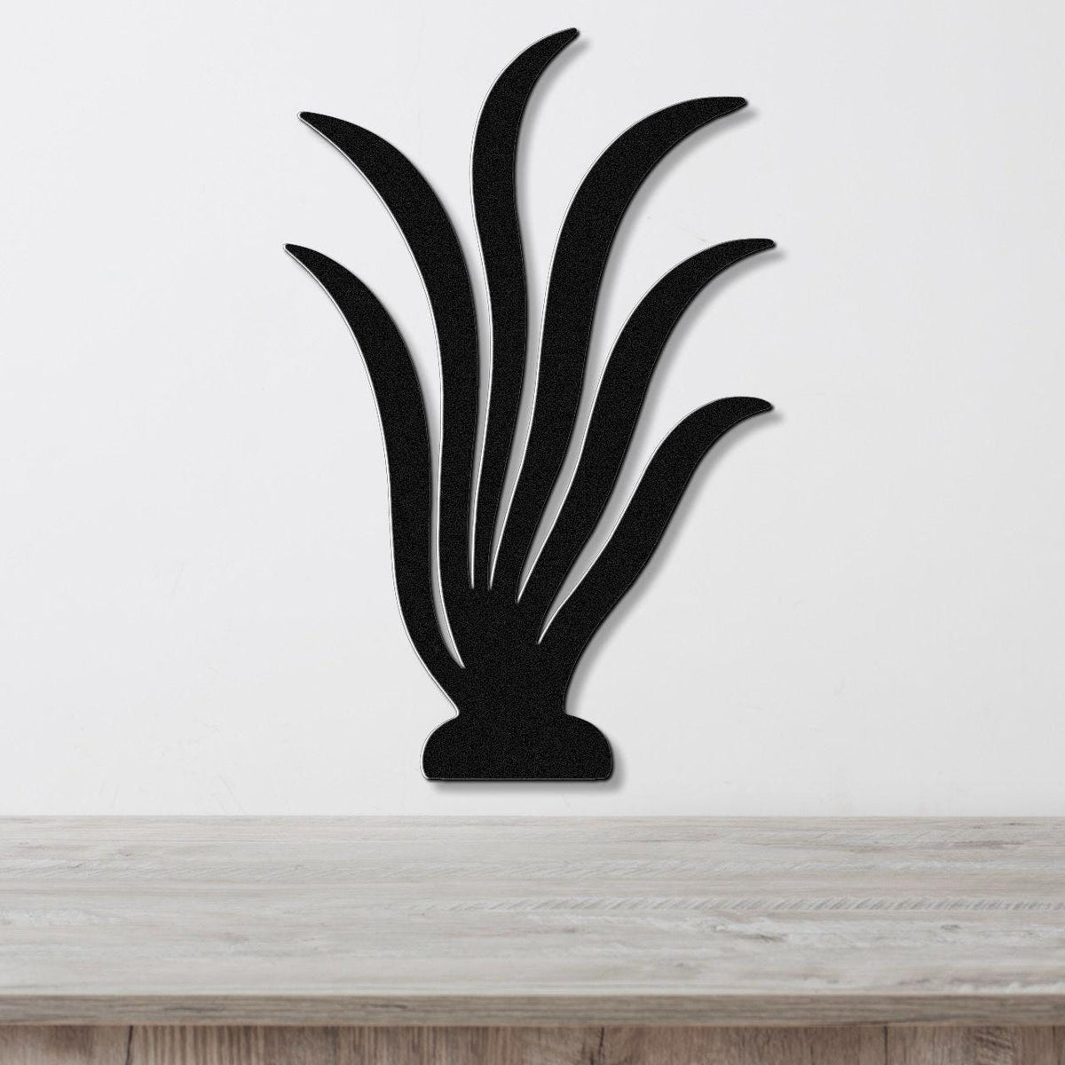 Black metal wall art cutout of a kelp or seaweed plant with flowing fronds, mounted on a white wall above a light wood surface.