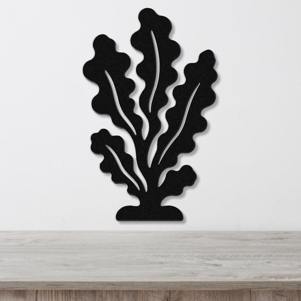 Black metal wall art in a kelp or seaweed silhouette with internal cutout veins, mounted on a white wall above a light wood surface.