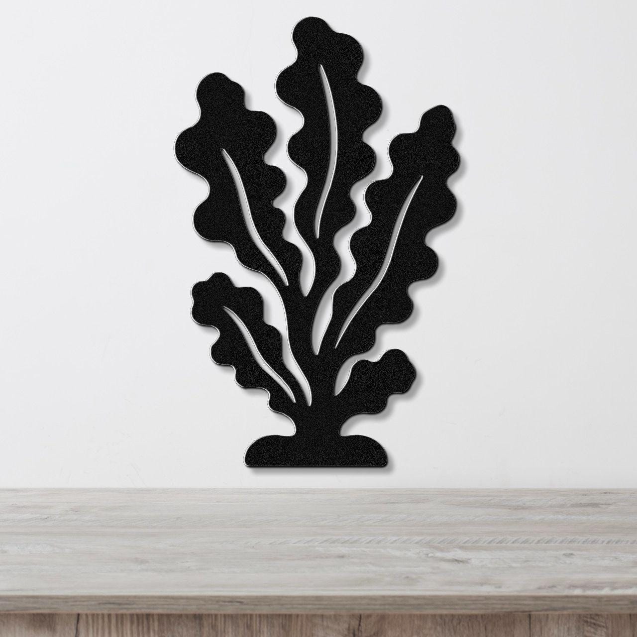 Black metal wall art in a kelp or seaweed silhouette with internal cutout veins, mounted on a white wall above a light wood surface.