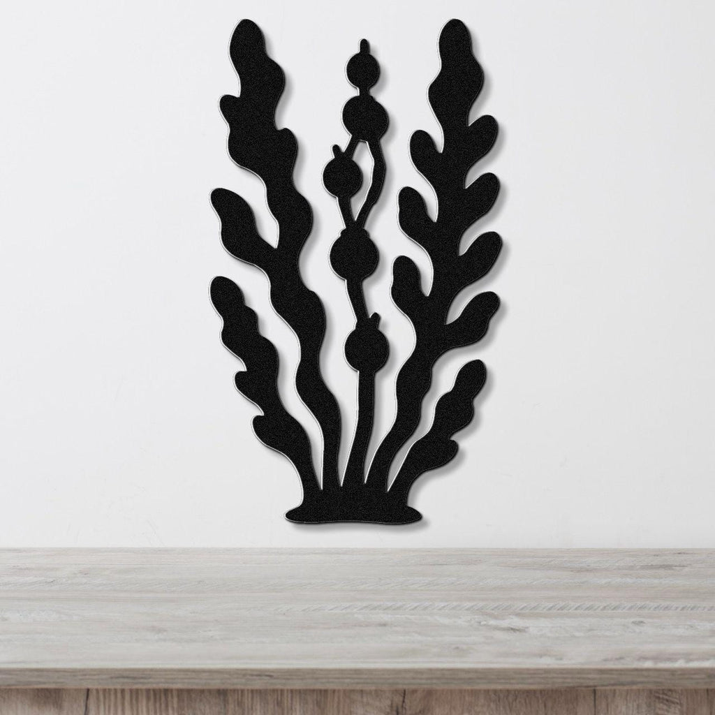 Black metal wall art cutout of kelp and seaweed with rounded fronds and bulbous stems, mounted on a white wall above a light wood surface.