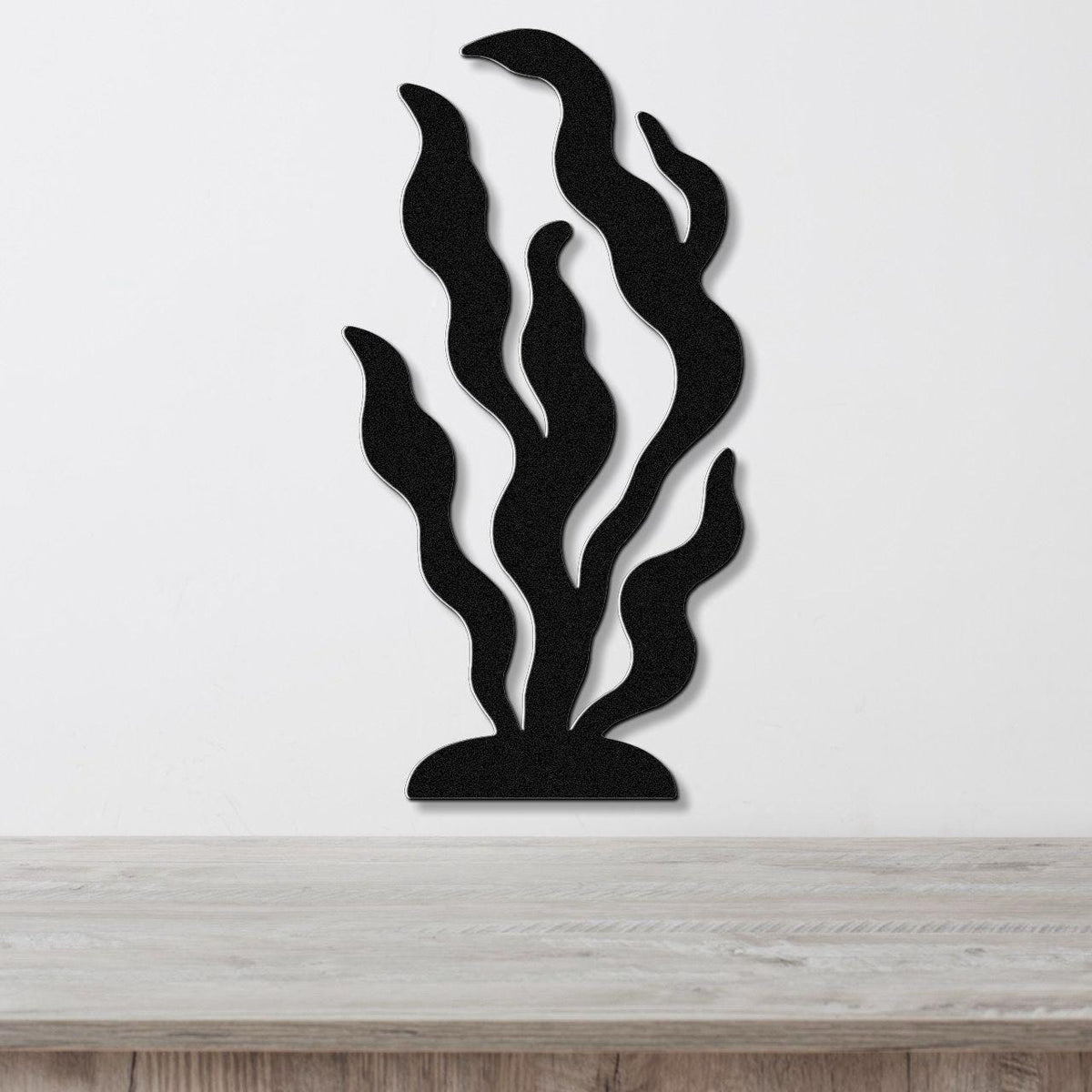 Black metal wall art in a kelp or seaweed silhouette design with layered, wavy fronds, mounted on a white wall above a light wood surface.