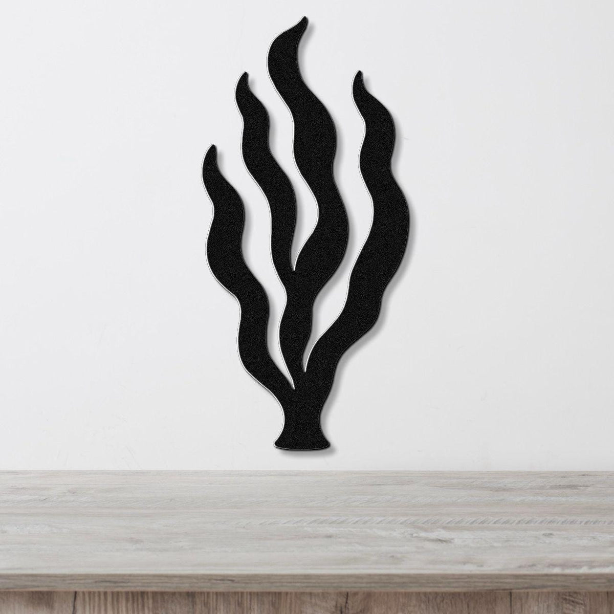Black metal wall art in a kelp seaweed silhouette with layered fronds, mounted on a white wall above a light wood surface.