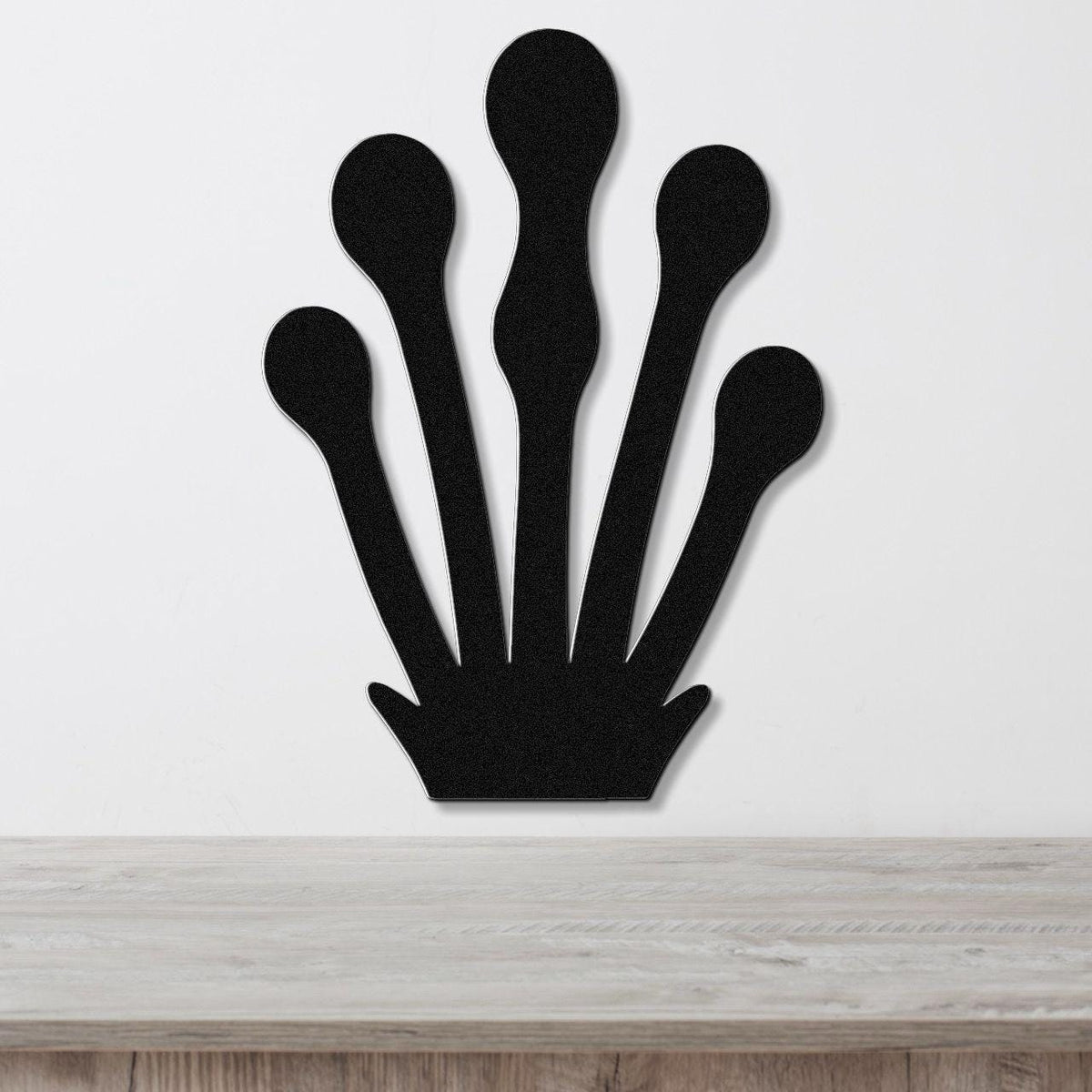 Black metal wall art in a kelp or seaweed silhouette design with five elongated fronds, mounted on a white wall above a light wood surface.