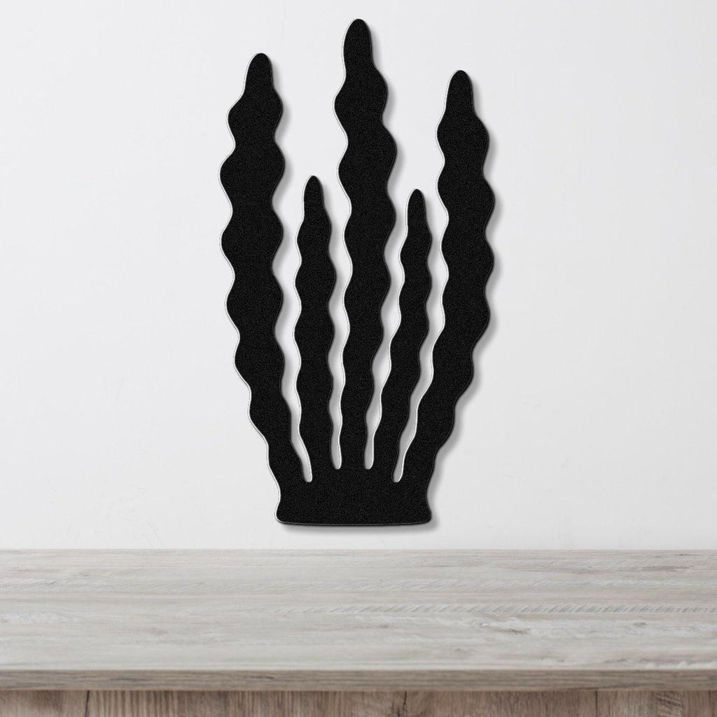 Black metal wall art with a kelp and seaweed silhouette design, featuring wavy cutouts, mounted on a white wall above a light wood surface.
