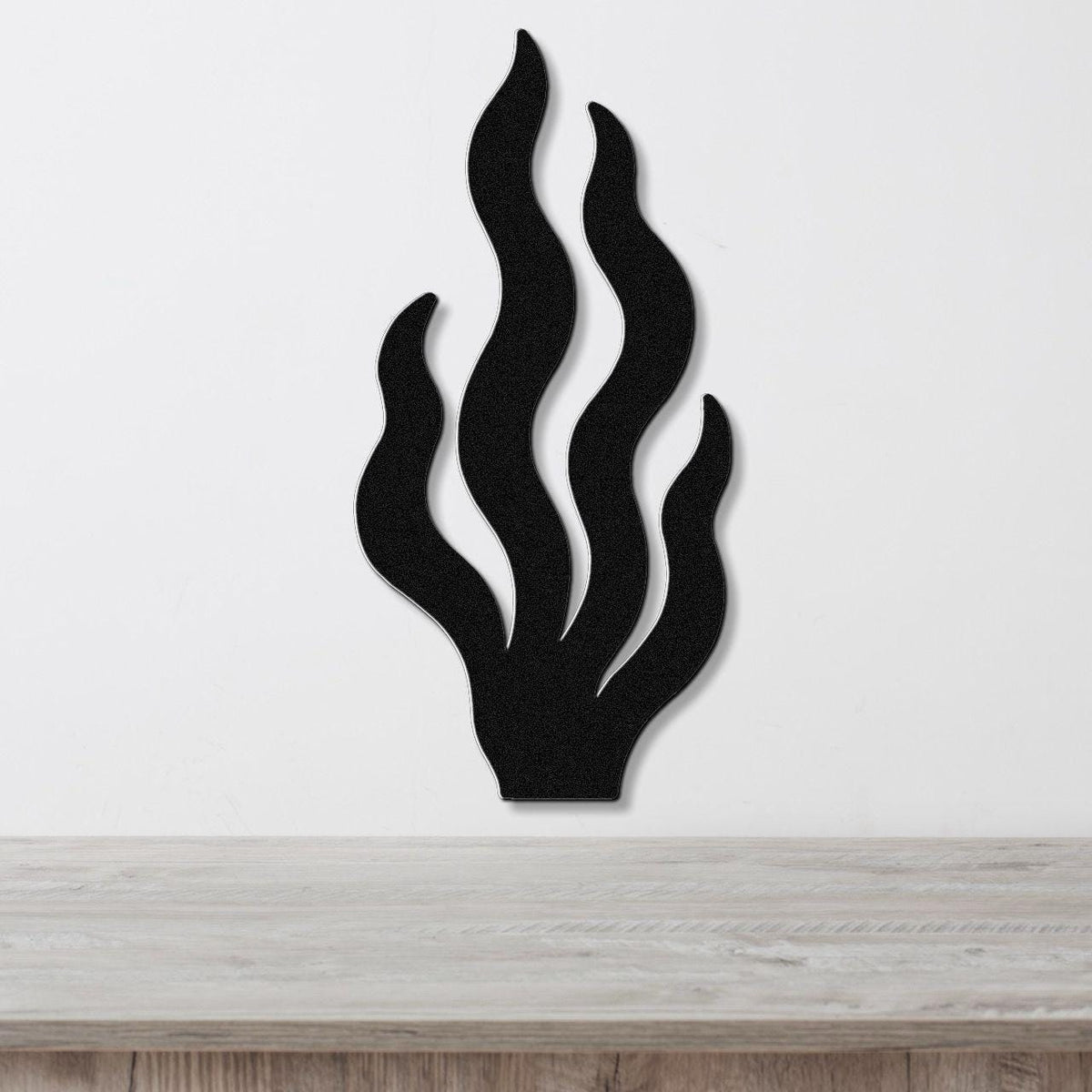 Black metal wall art in a kelp or seaweed silhouette with layered, flowing fronds, mounted on a white wall above a light wood surface.