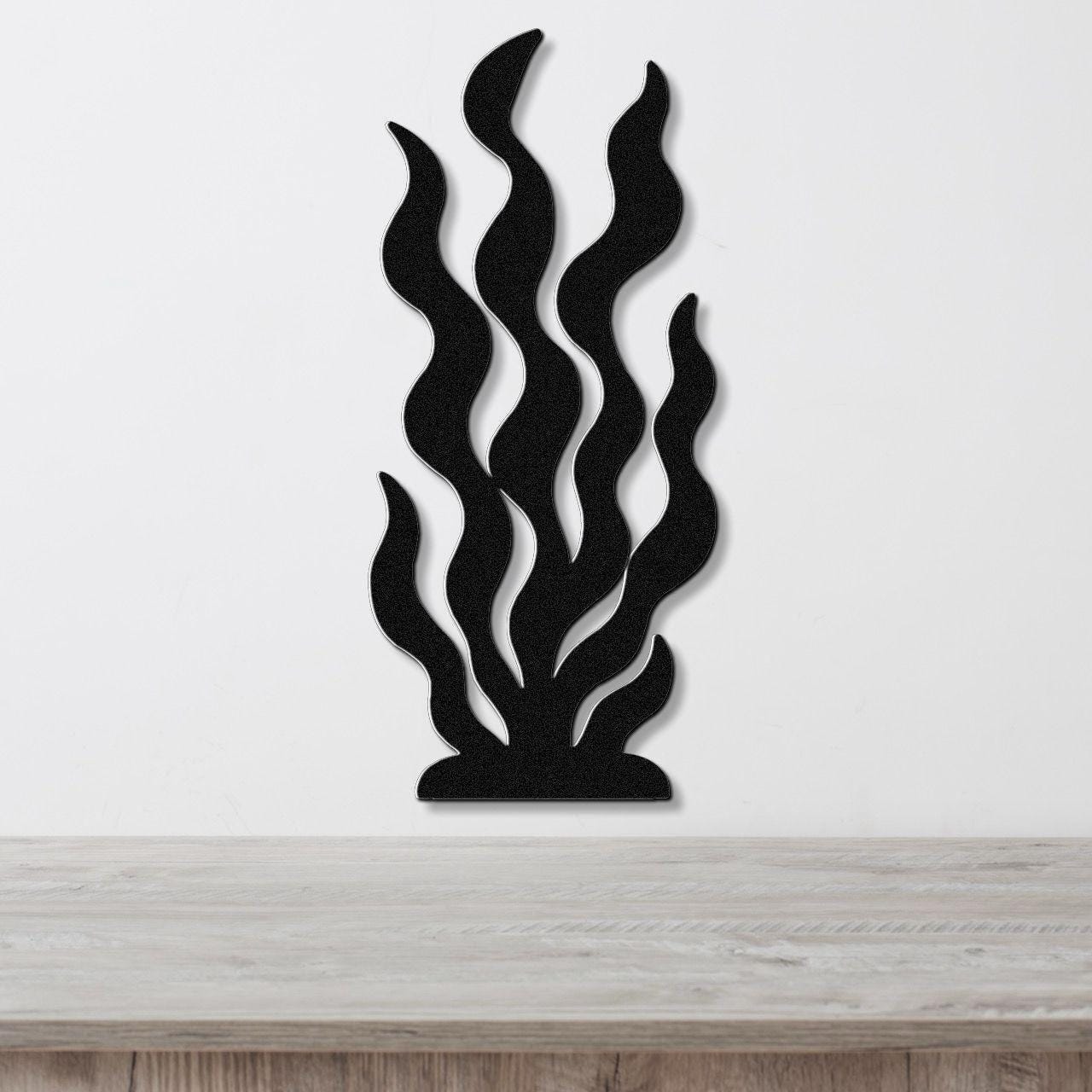 Black metal wall art in a seaweed or kelp silhouette design with layered, wavy cutouts, mounted on a white wall above a light wood surface.